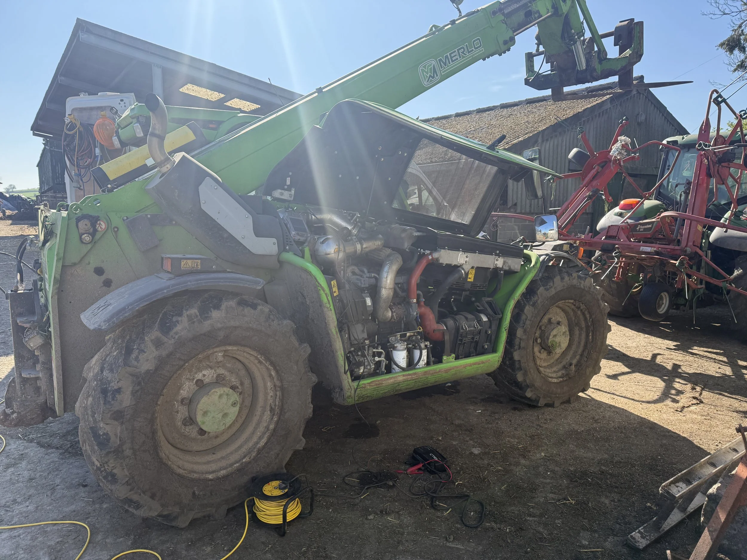 A green Merlo crane attached to an all-terrain vehicle is lifting a large container or bin in a farm or rural setting. The scene includes gardening or farming tools, extension cords, machinery, and a barn in the background under a clear blue sky.