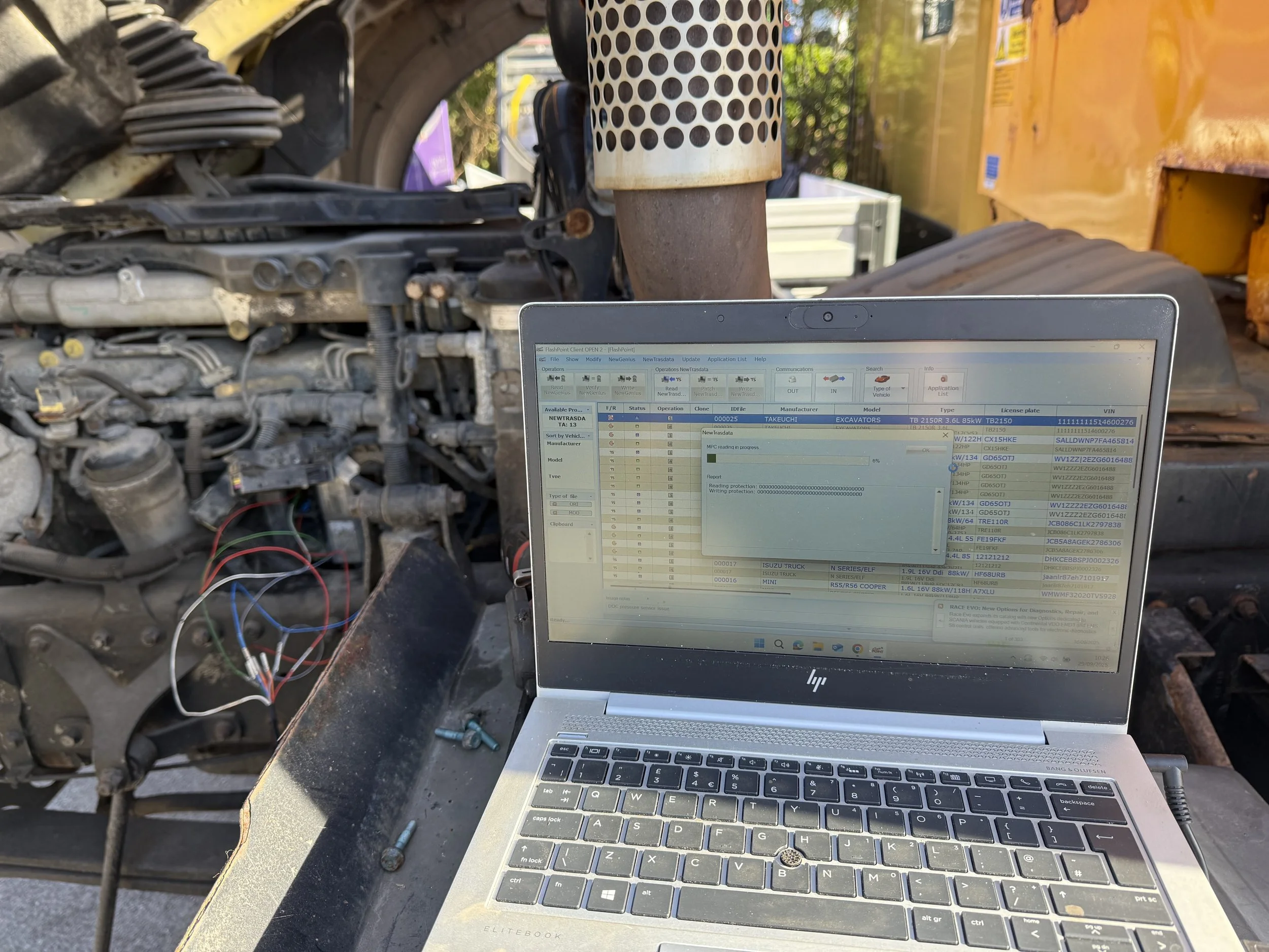 This image shows a laptop computer on a work surface with a screen displaying data and information. Behind the laptop, there are various mechanical parts and components, possibly from a piece of heavy machinery or an engine, with wires and hoses visi