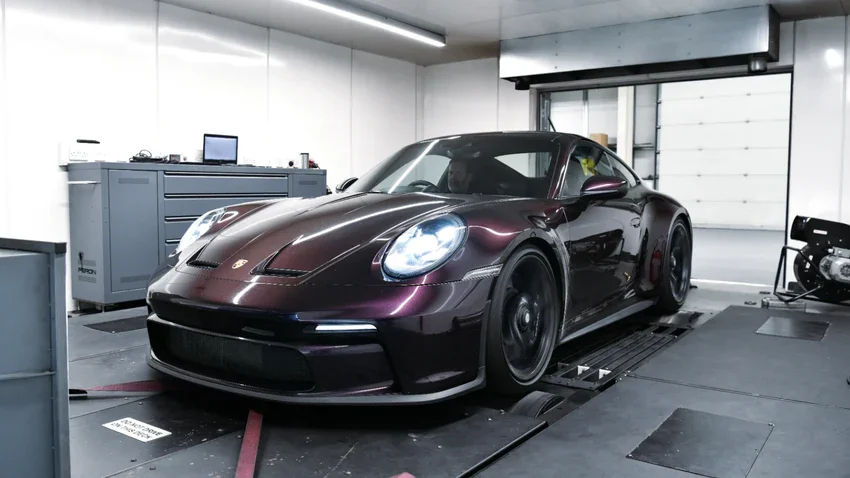 A dark purple Porsche sports car inside a garage or testing station, with equipment and a computer on a workbench in the background.