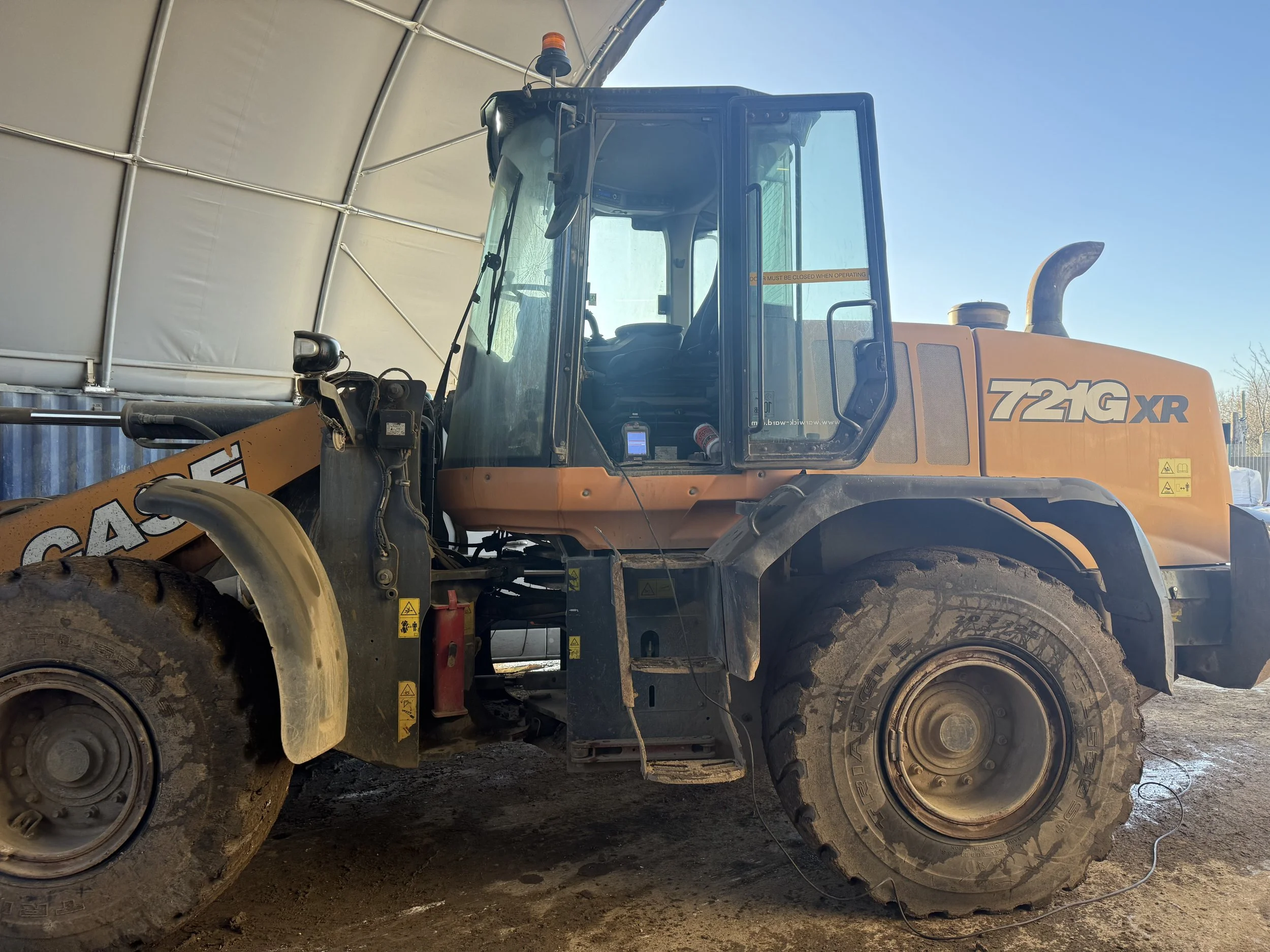 A large construction or industrial vehicle with the model number 721G XR, parked inside a covered structure with a blue sky visible overhead.