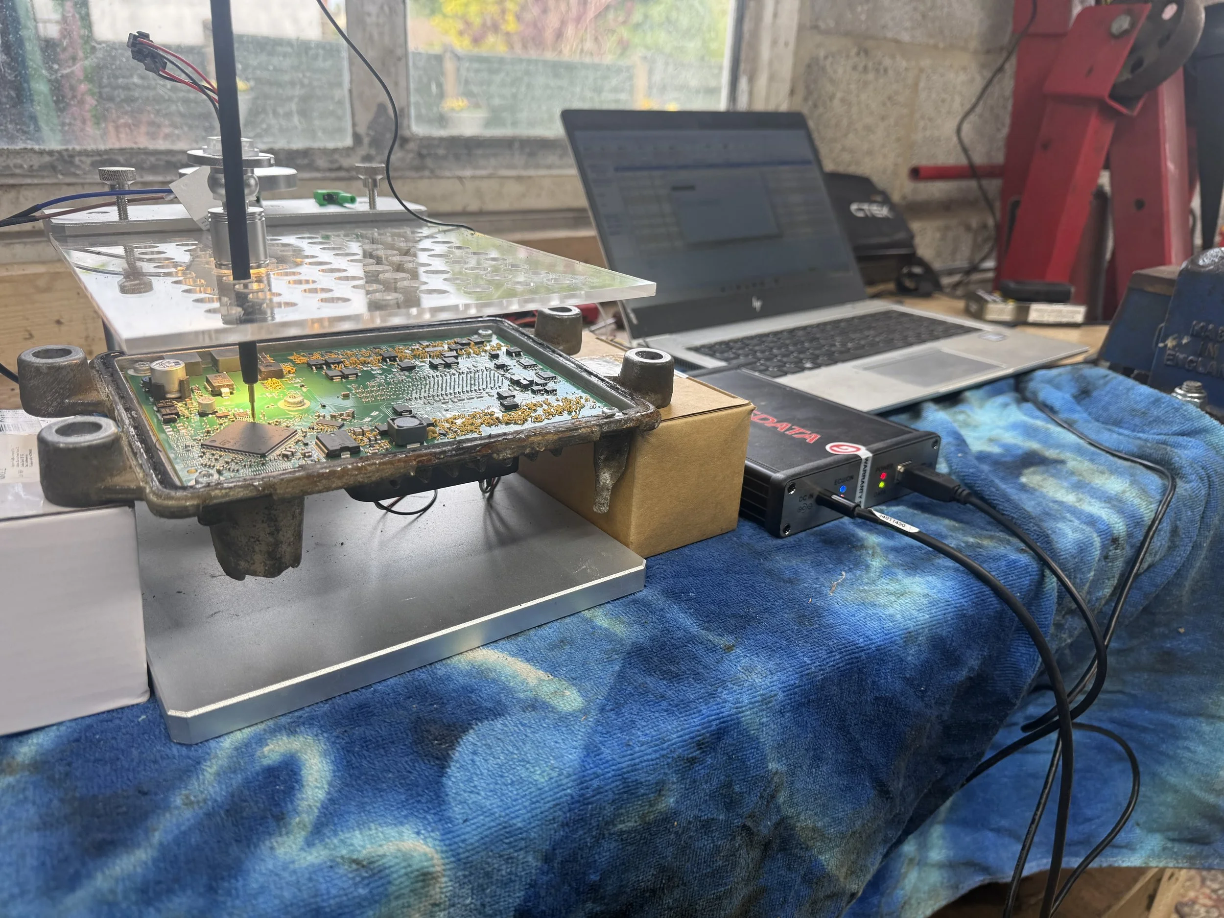Inside a workshop, a workbench holds a disassembled electronic circuit board, a laptop, and various tools. The background shows a window with a view of trees.