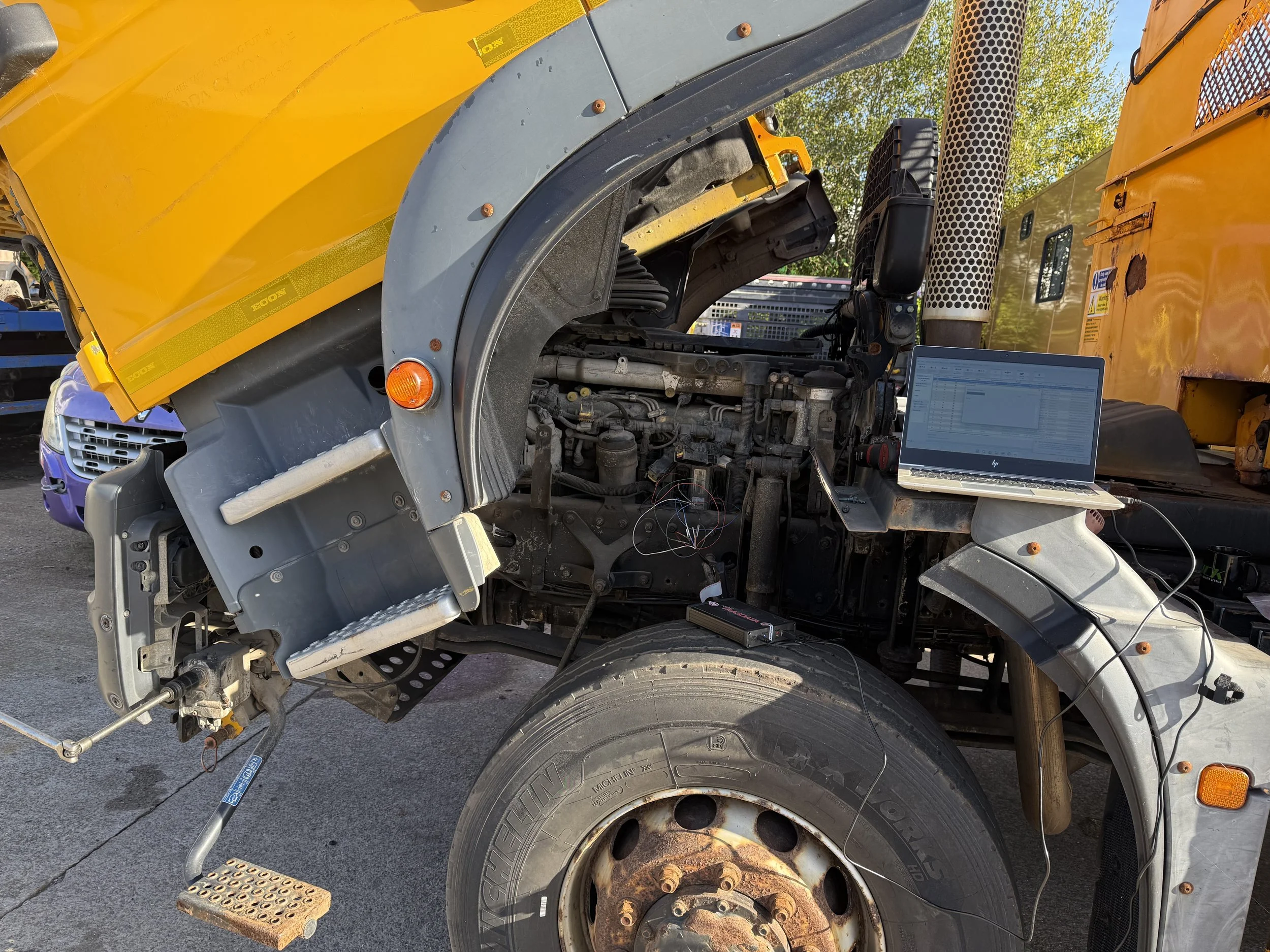 Partial view of a yellow and black snomobile or heavy machinery with exposed mechanical parts, a laptop computer on the side, a tire, and some tools nearby, indicating maintenance or repair work.