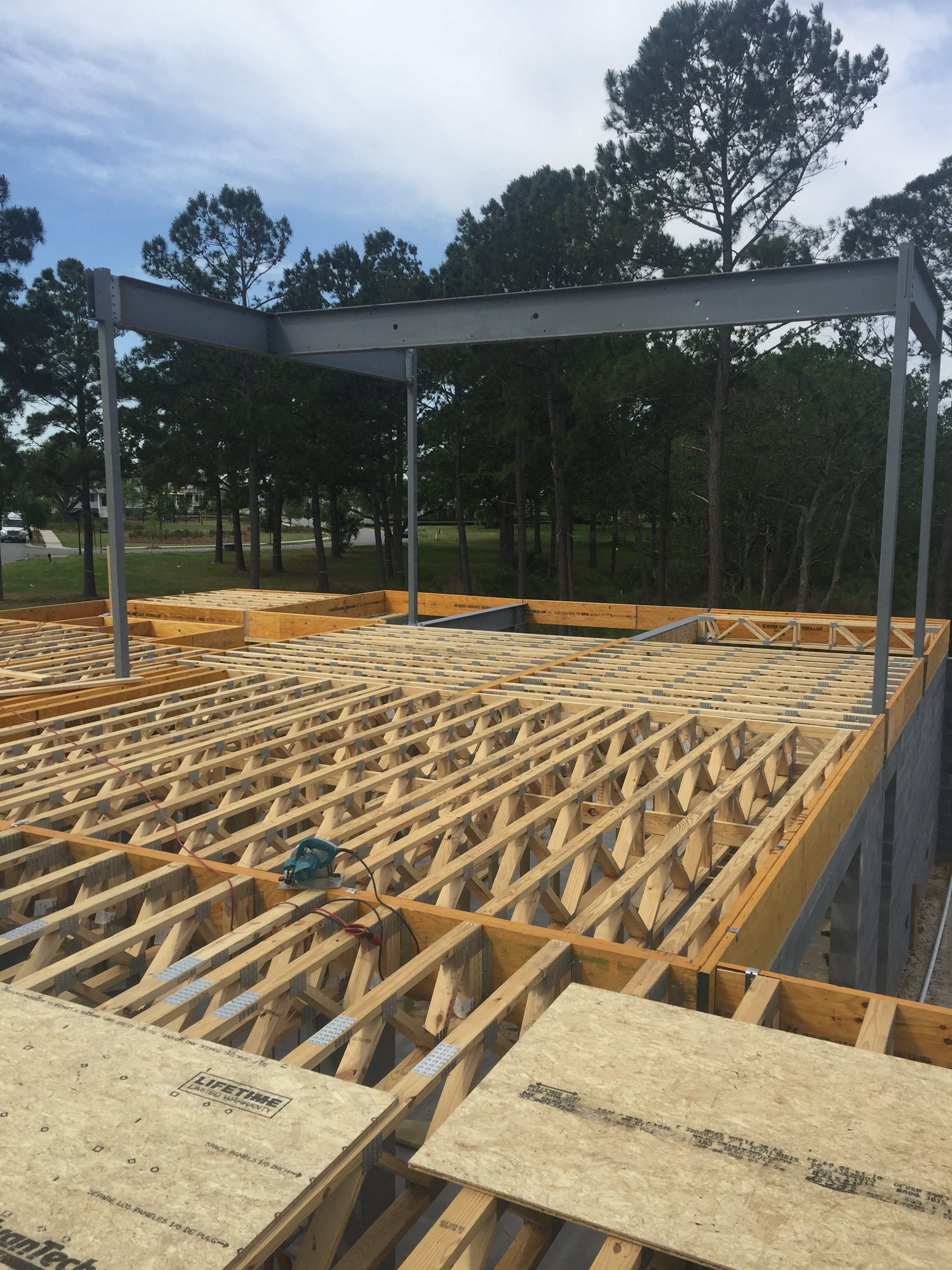 Construction site with wooden framing and steel beam structure, set against a background of pine trees and a partly cloudy sky.
