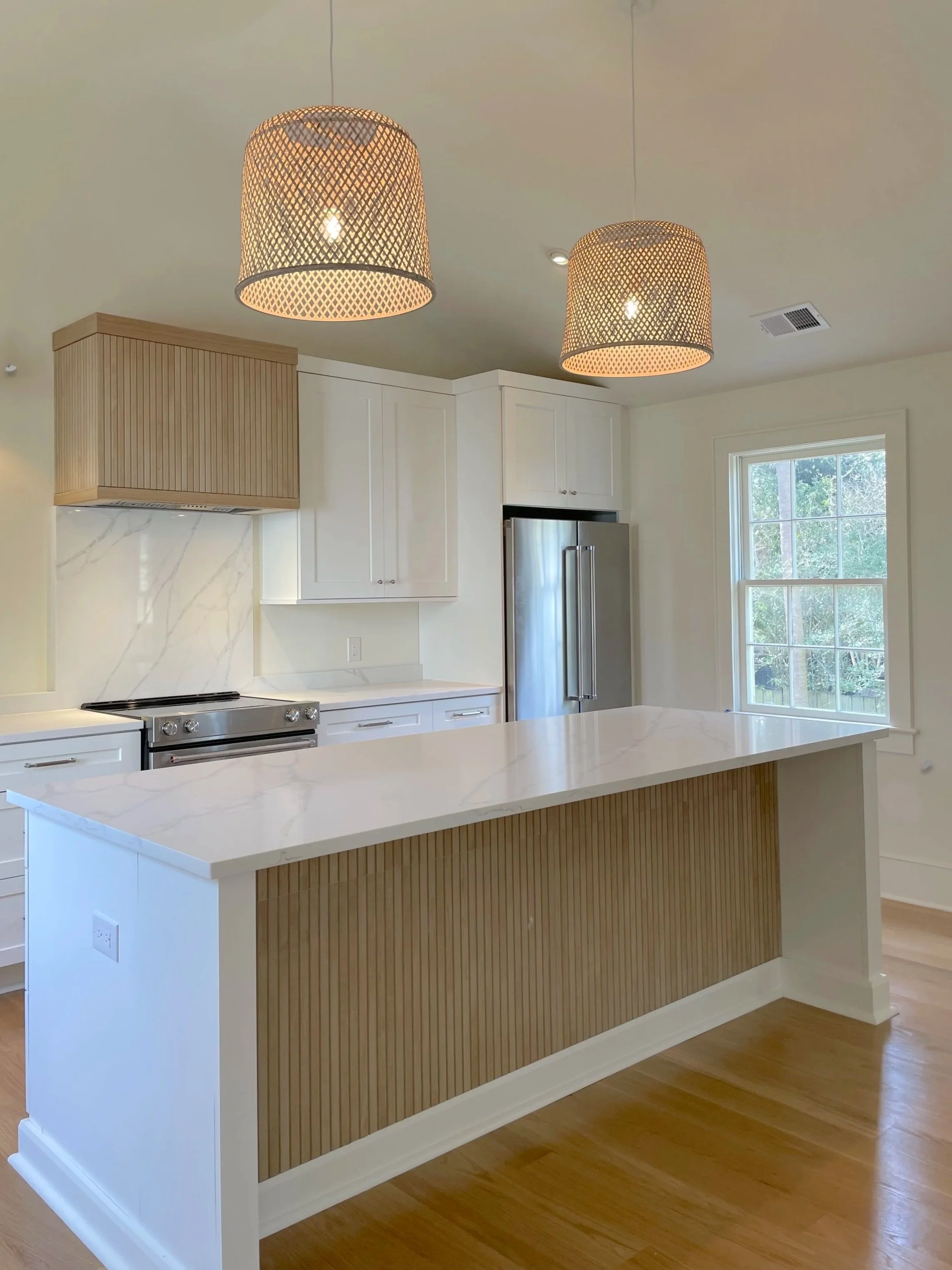 Modern kitchen with white cabinets, a marble backsplash, stainless steel refrigerator, and a large island with wooden paneling. Two woven pendant lights hang from the ceiling, and a large window provides natural light.