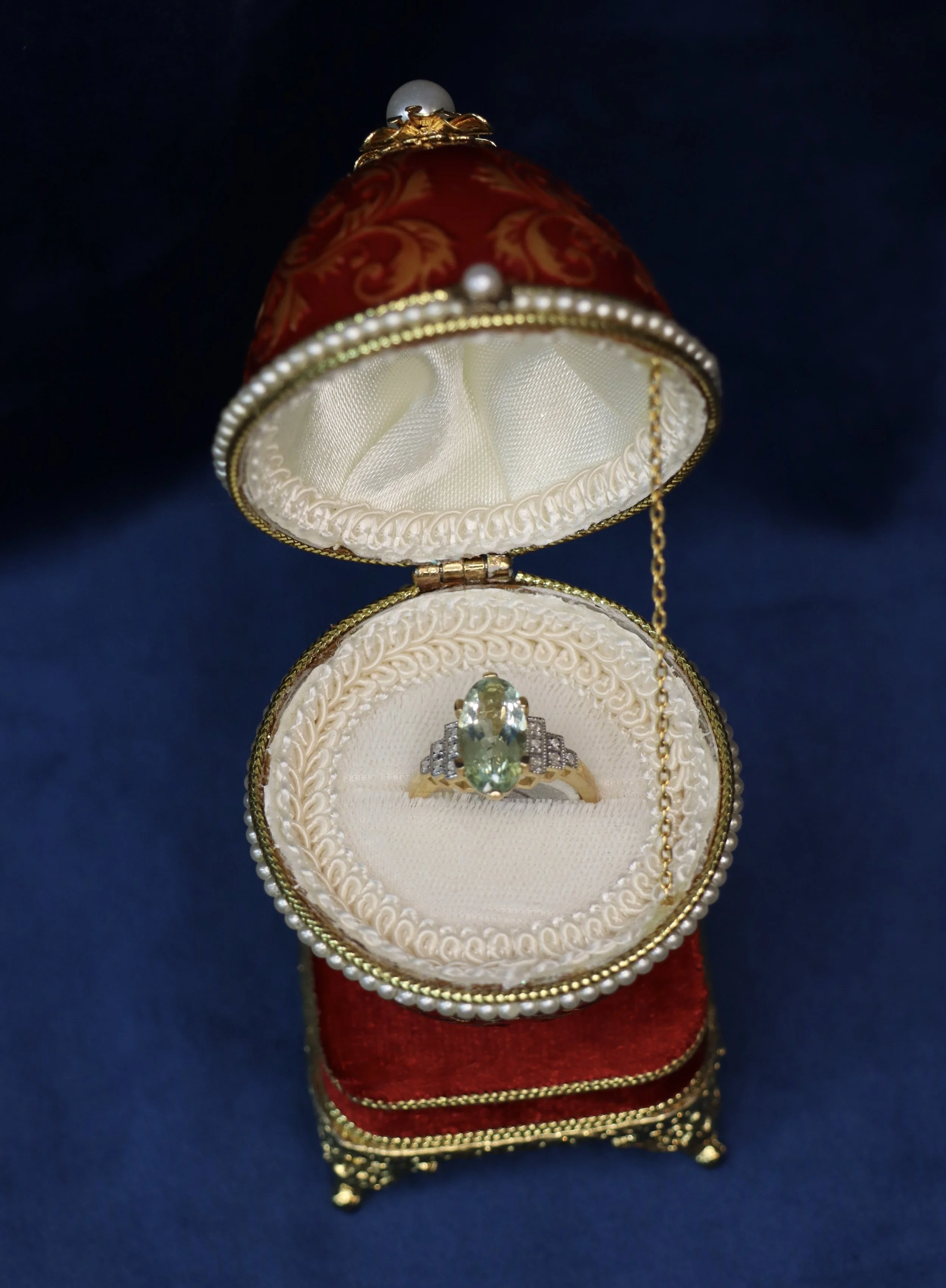 A jewelry ring with a large oval green gemstone, surrounded by smaller clear stones, displayed in an ornate round jewelry box with a white interior, gold trim, and a chain attached, set against a dark background.
