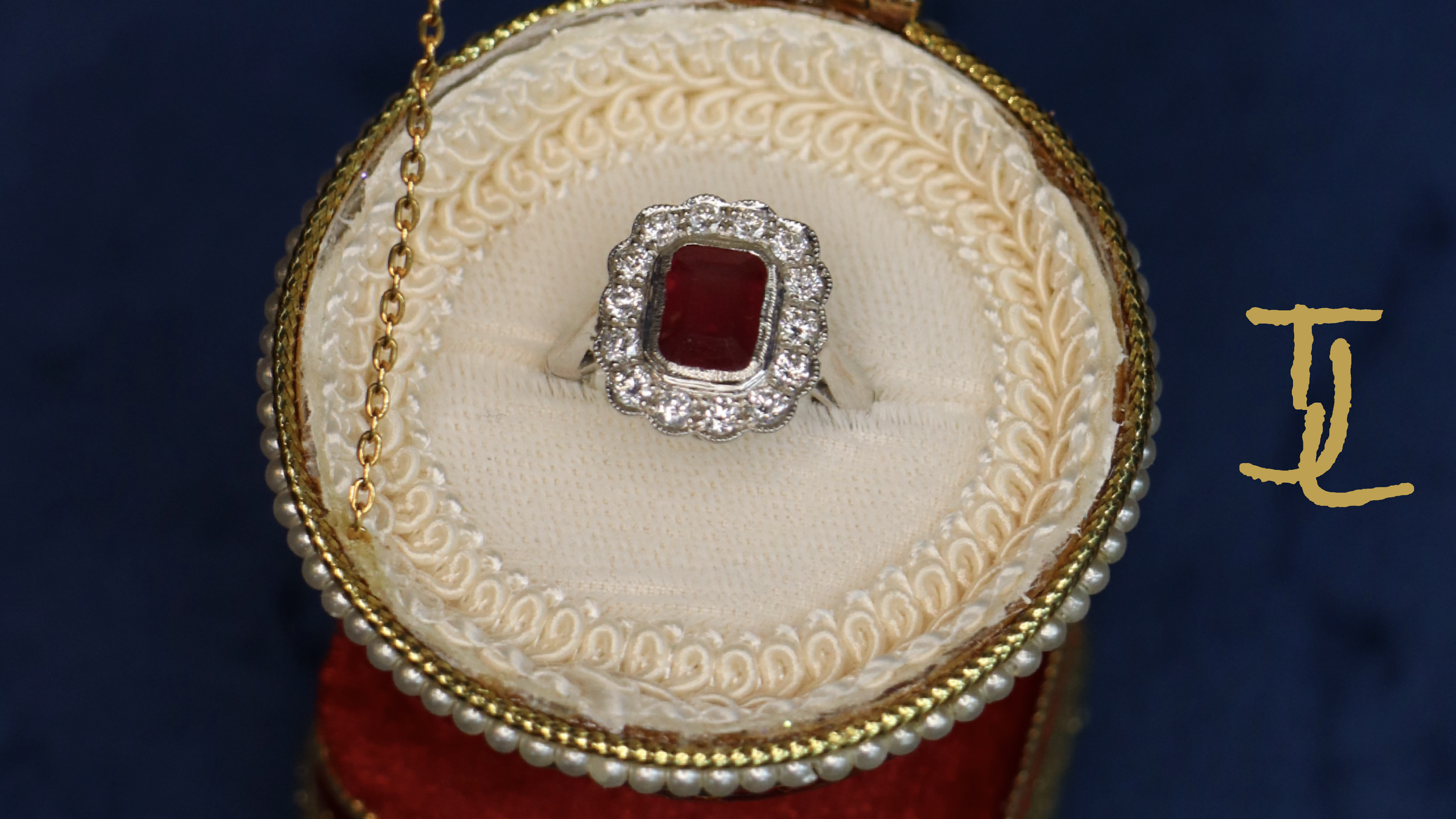 A platinum ring with a large rectangular ruby gemstone surrounded by small diamonds, displayed on a decorative cushion inside a jewelry box with gold trim and pearl accents.