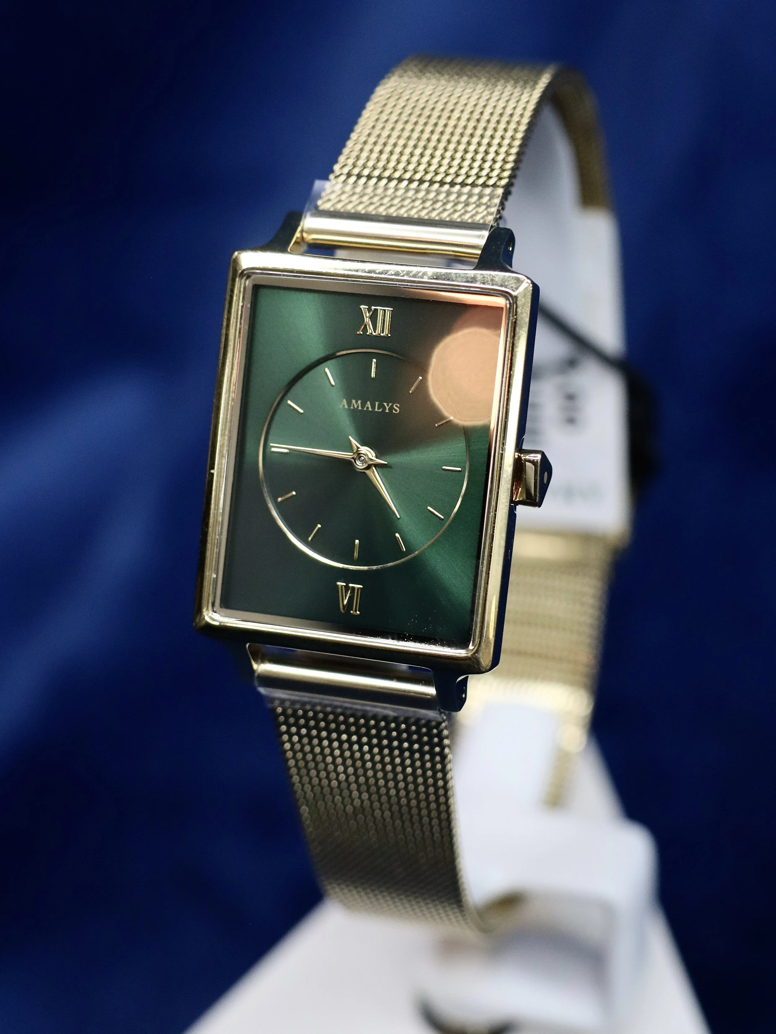 Close-up of a rectangular wristwatch with a gold mesh strap and a green dial, displaying the time with gold hands, set against a dark blue background.