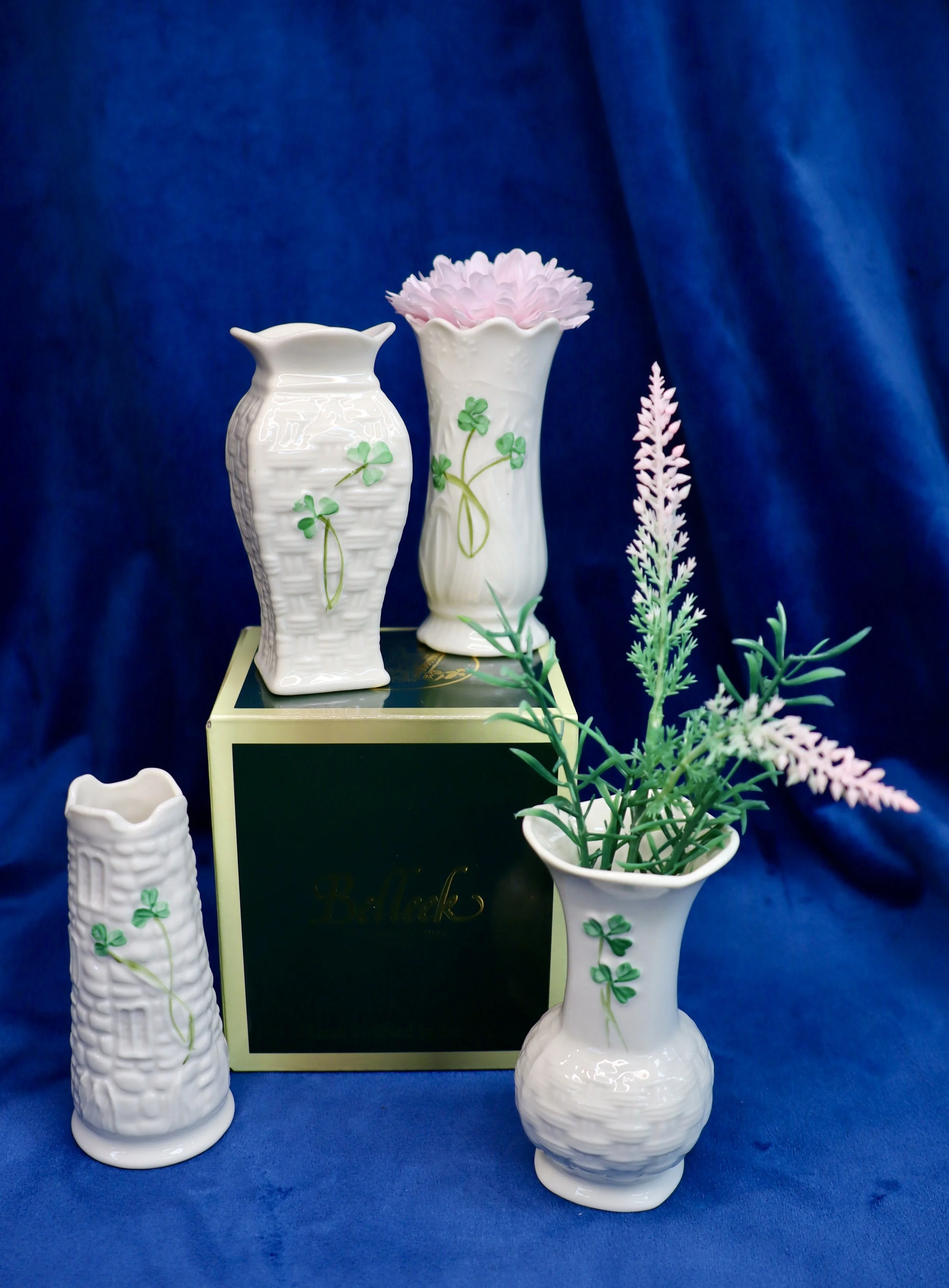Collection of white ceramic vases with shamrock and clover decoration, greenery and artificial flowers, set against a dark blue background.
