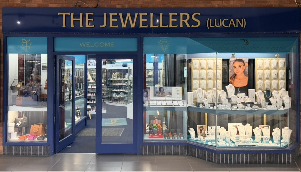 Jewelry store storefront named 'The Jewellers' with display windows showcasing necklaces, rings, and bracelets, and glass entrance door with 'Welcome' sign.