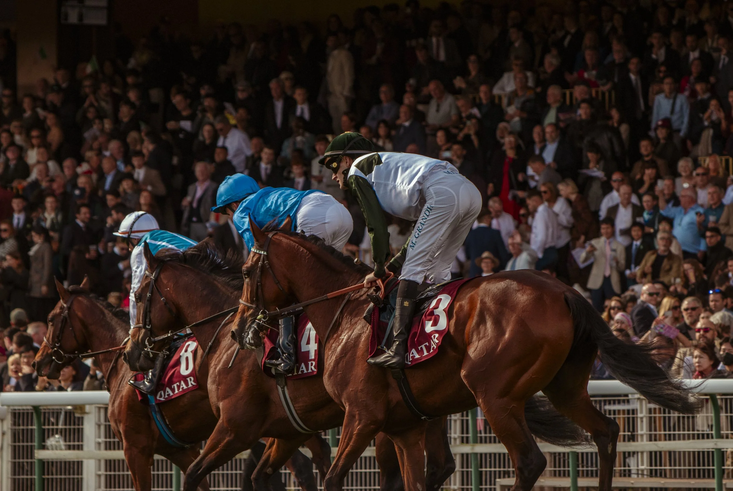 Jockeys riding racehorses during a horse race with a large crowd of spectators in the background.
