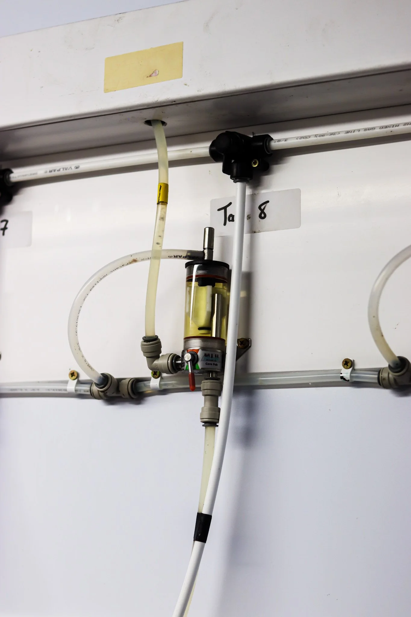 Close-up of a beer tap setup with pipes, valves, and a pressure gauge mounted on a white wall in an industrial or laboratory environment. Beer room