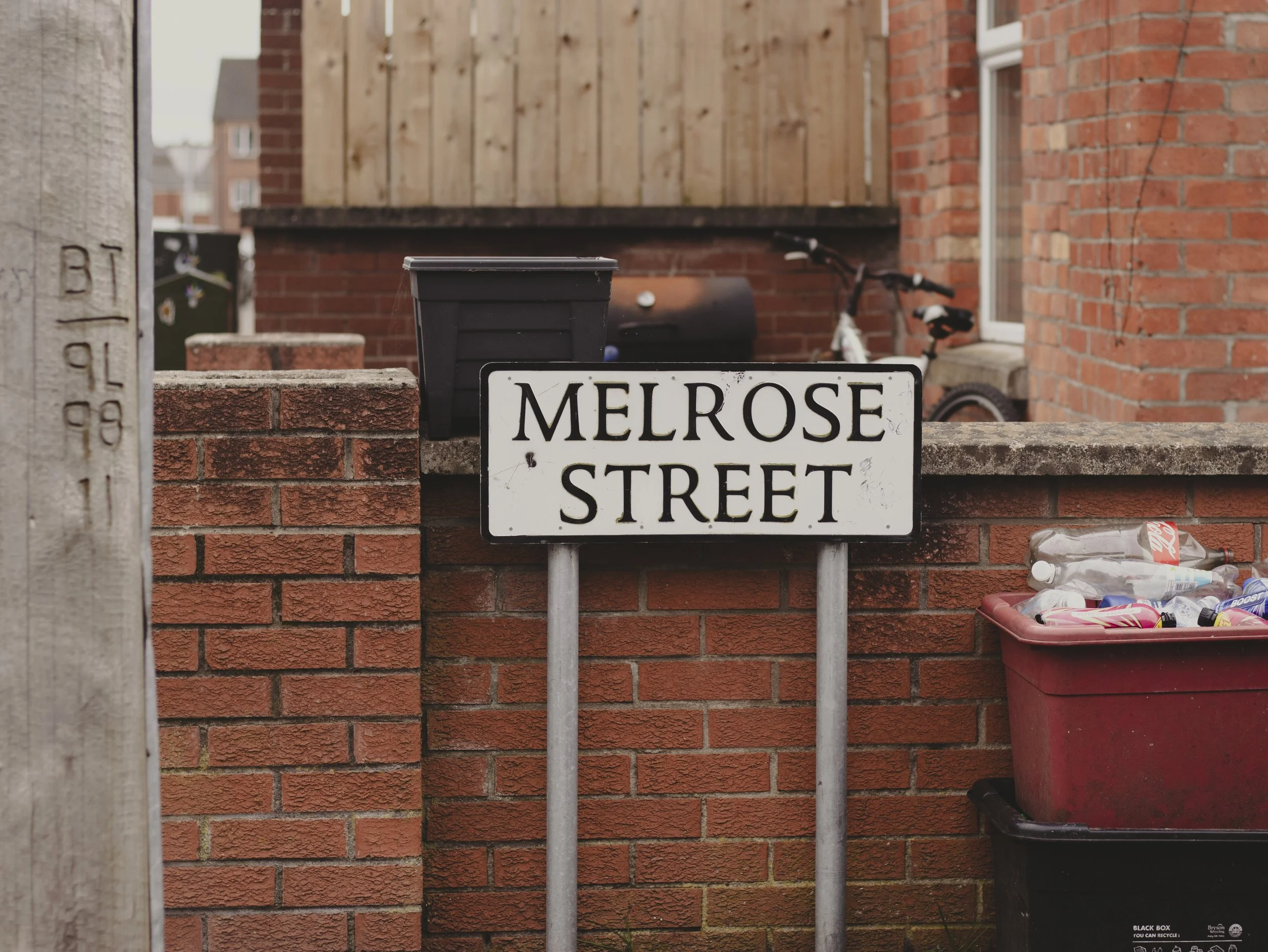 A street sign reading 'Melrose Street' in front of a brick wall, a black mailbox, a trash bin, and a bicycle partially visible behind the mailbox, with a red recycling bin containing plastic bottles. Belfast