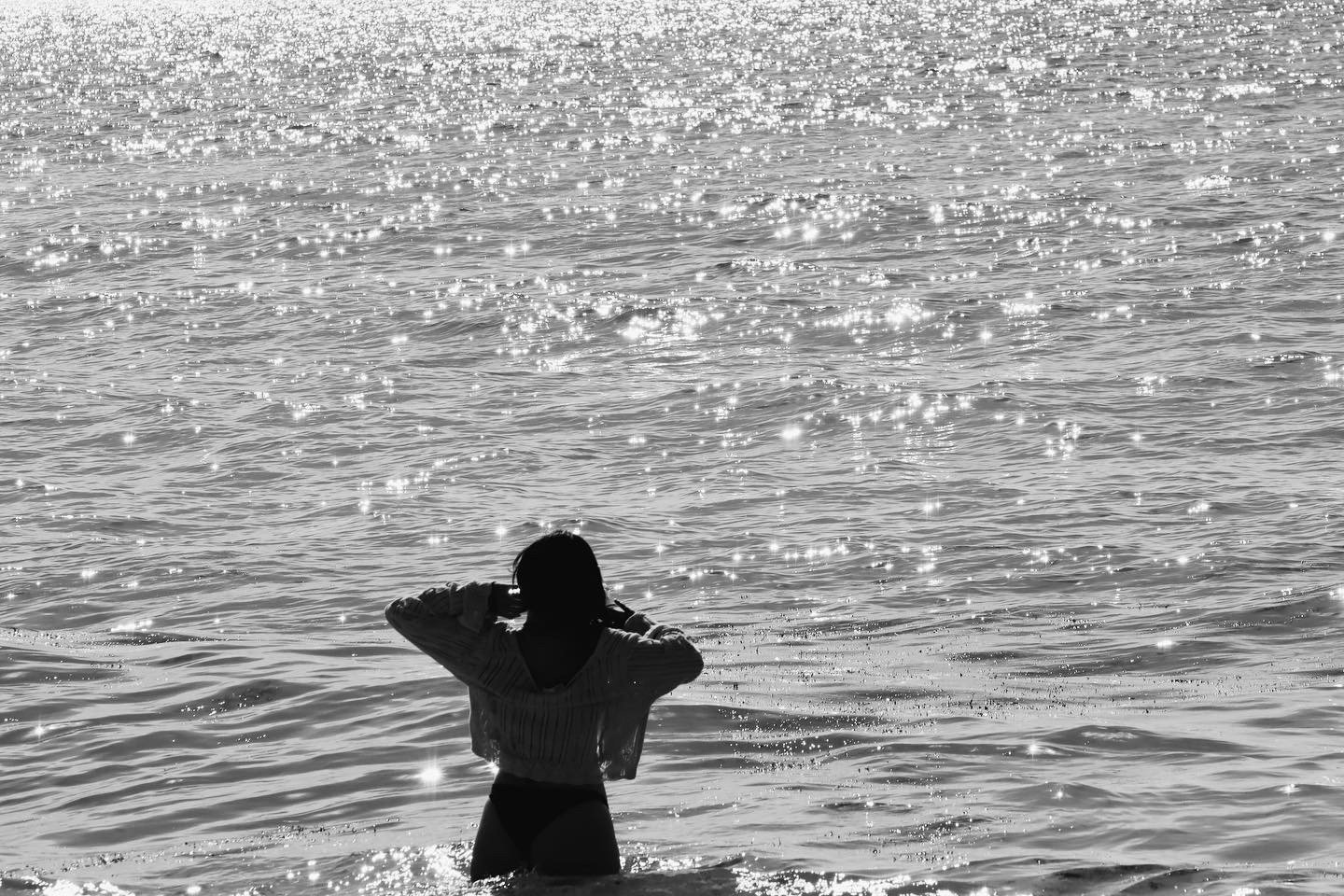 Silhouette of a woman standing in the water at the beach, facing away, with her arms raised to adjust her hair, sunlight reflecting on the water surface.