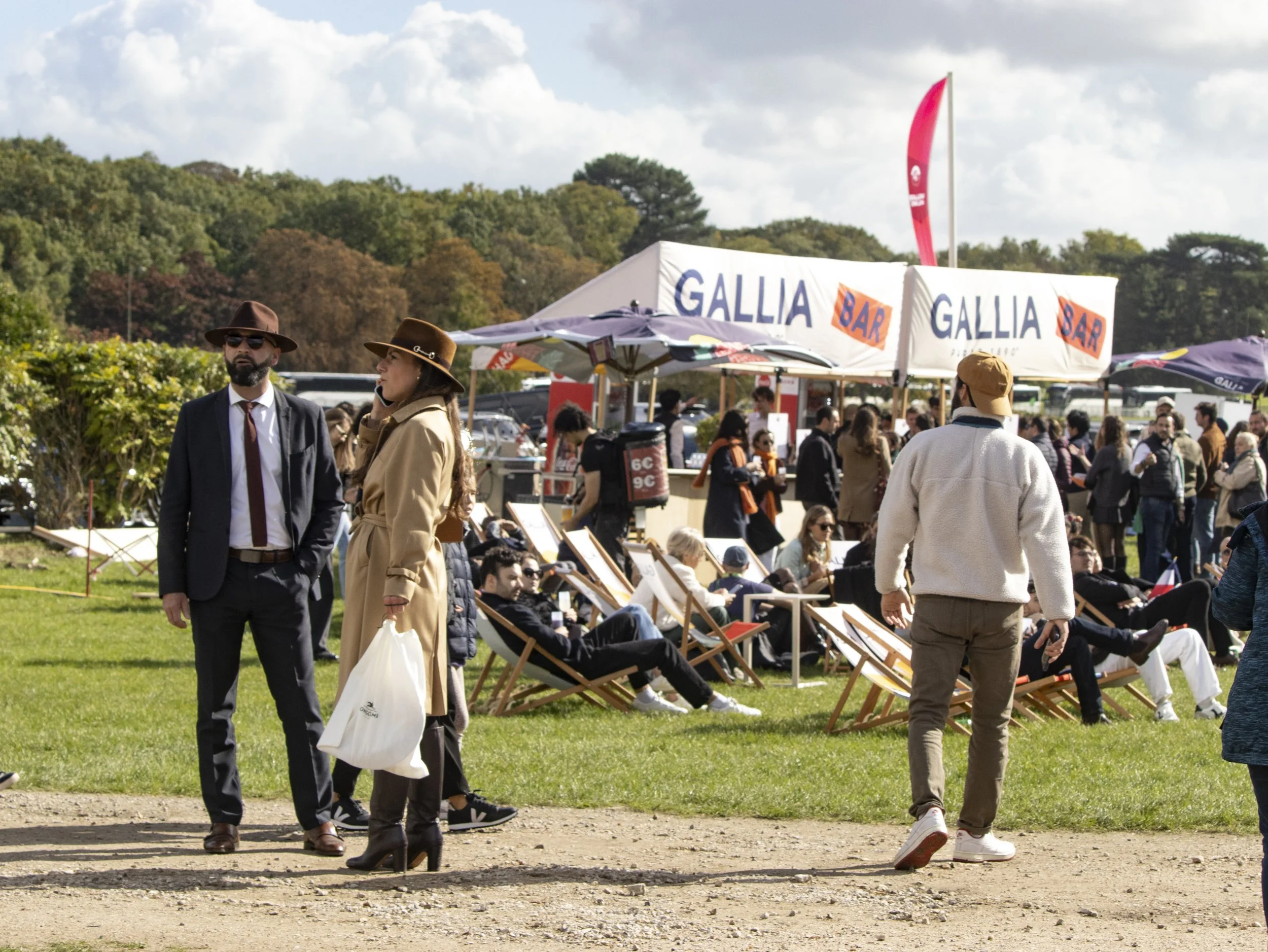 People walking and sitting at an outdoor event with tents and banners, including a large sign that reads 'GALLIA BAR' in a park with trees and a cloudy sky.