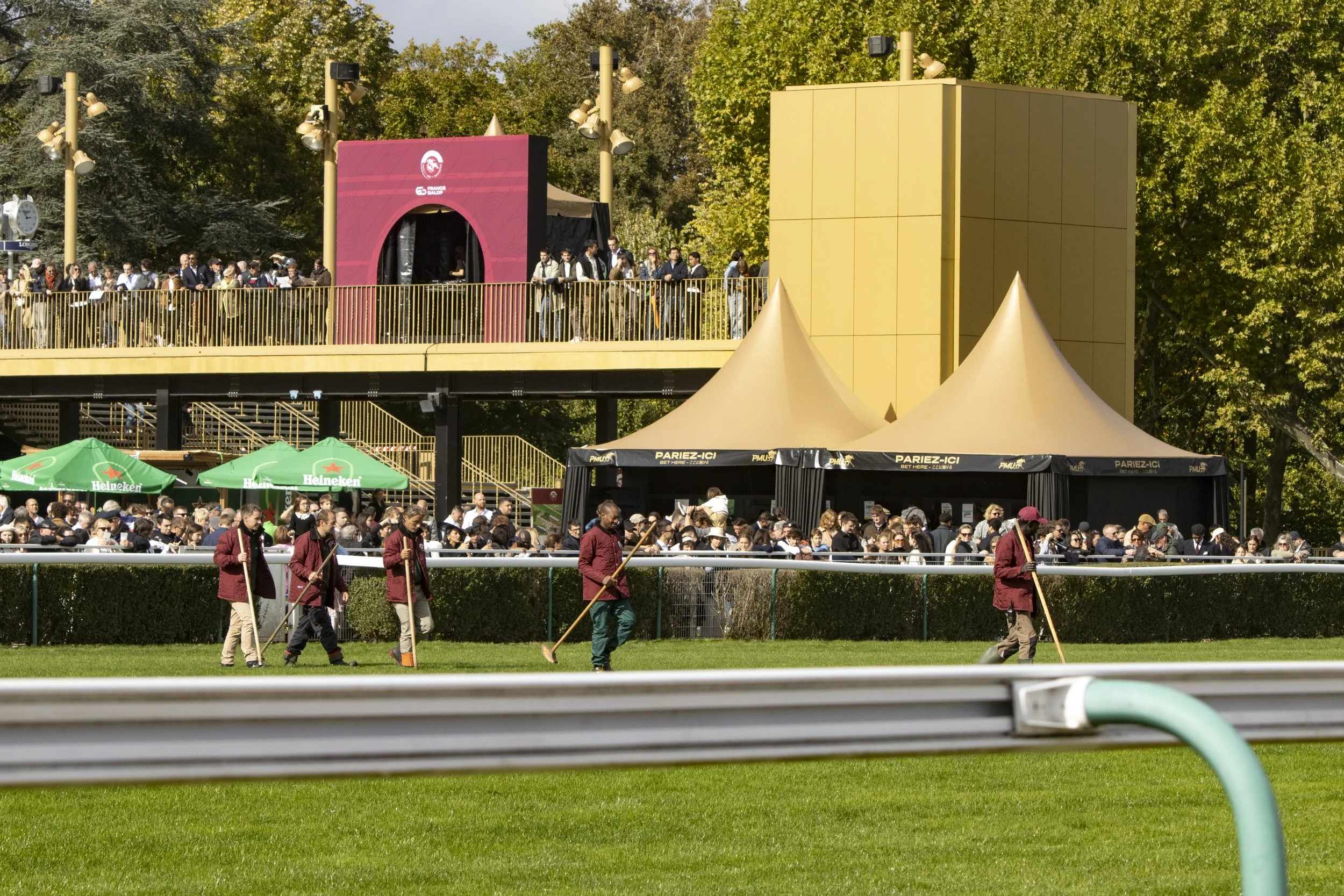 Crowd of people gathered on a balcony above a green field, with four workers in maroon uniforms working on the grass in the foreground. Large trees and tents are in the background.