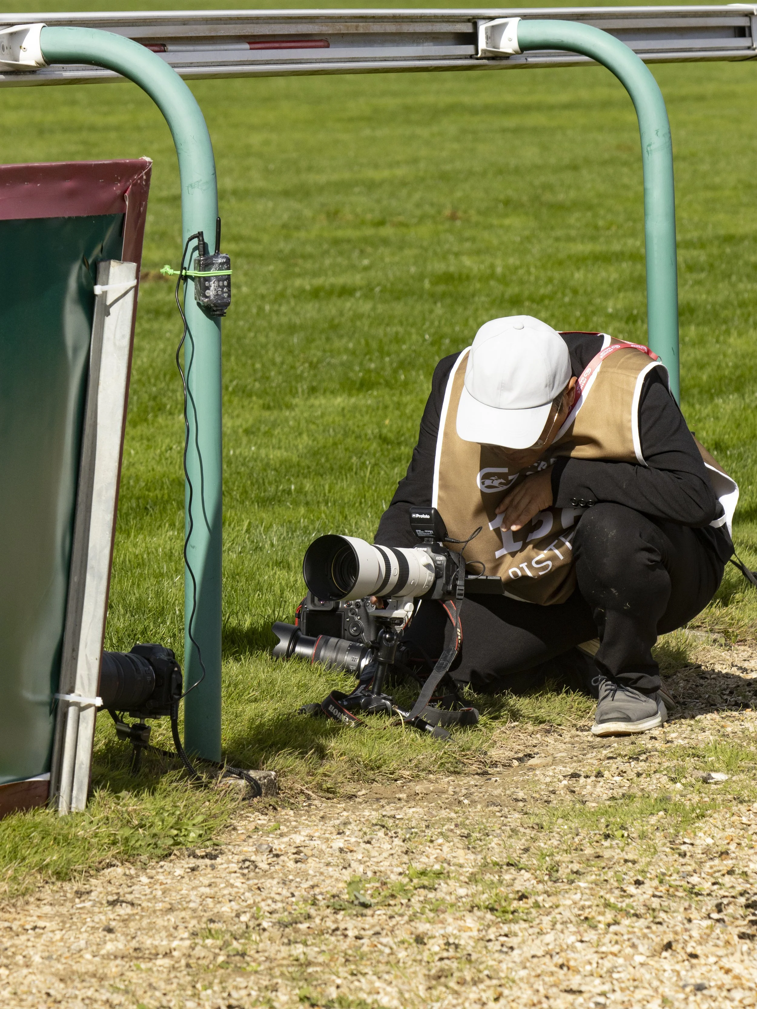 Photographer crouching on grass with professional camera, under a sports stadium canopy, taking pictures of an event.