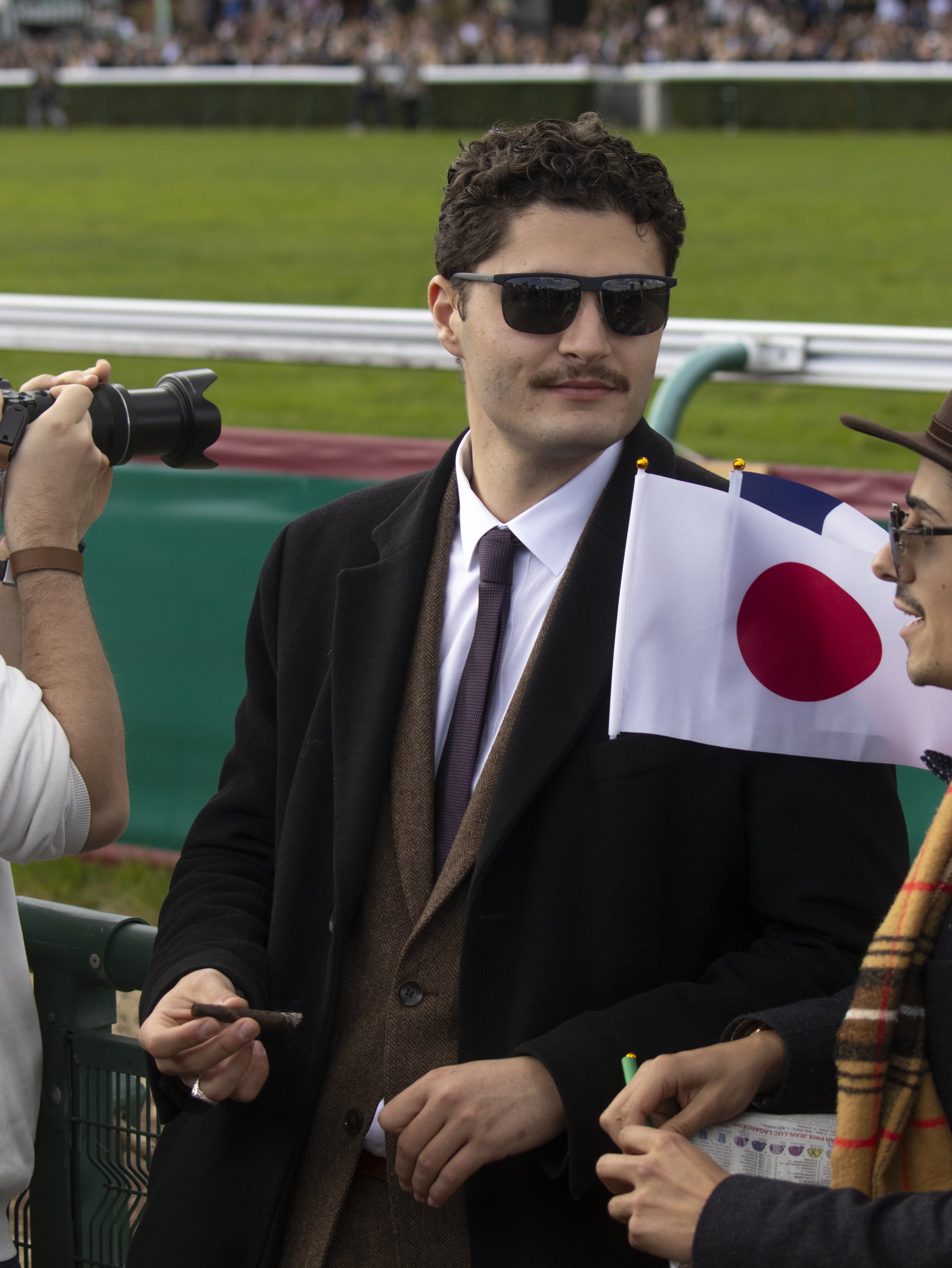 A man with curly hair and sunglasses wearing a suit and tie holding a small Japanese flag at a horse racing event, with other people nearby and a grassy racecourse in the background.