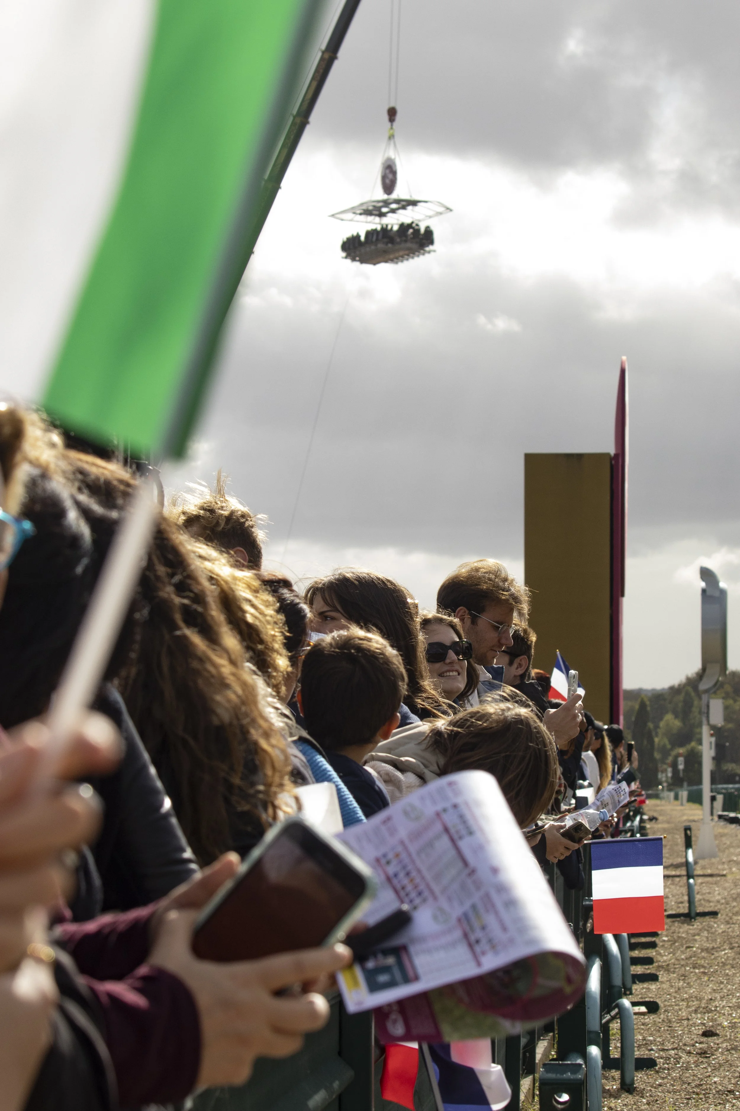 A crowd of people standing in a line outdoors, many holding papers or brochures, with flags visible in the background. Overcast sky with dark clouds.
