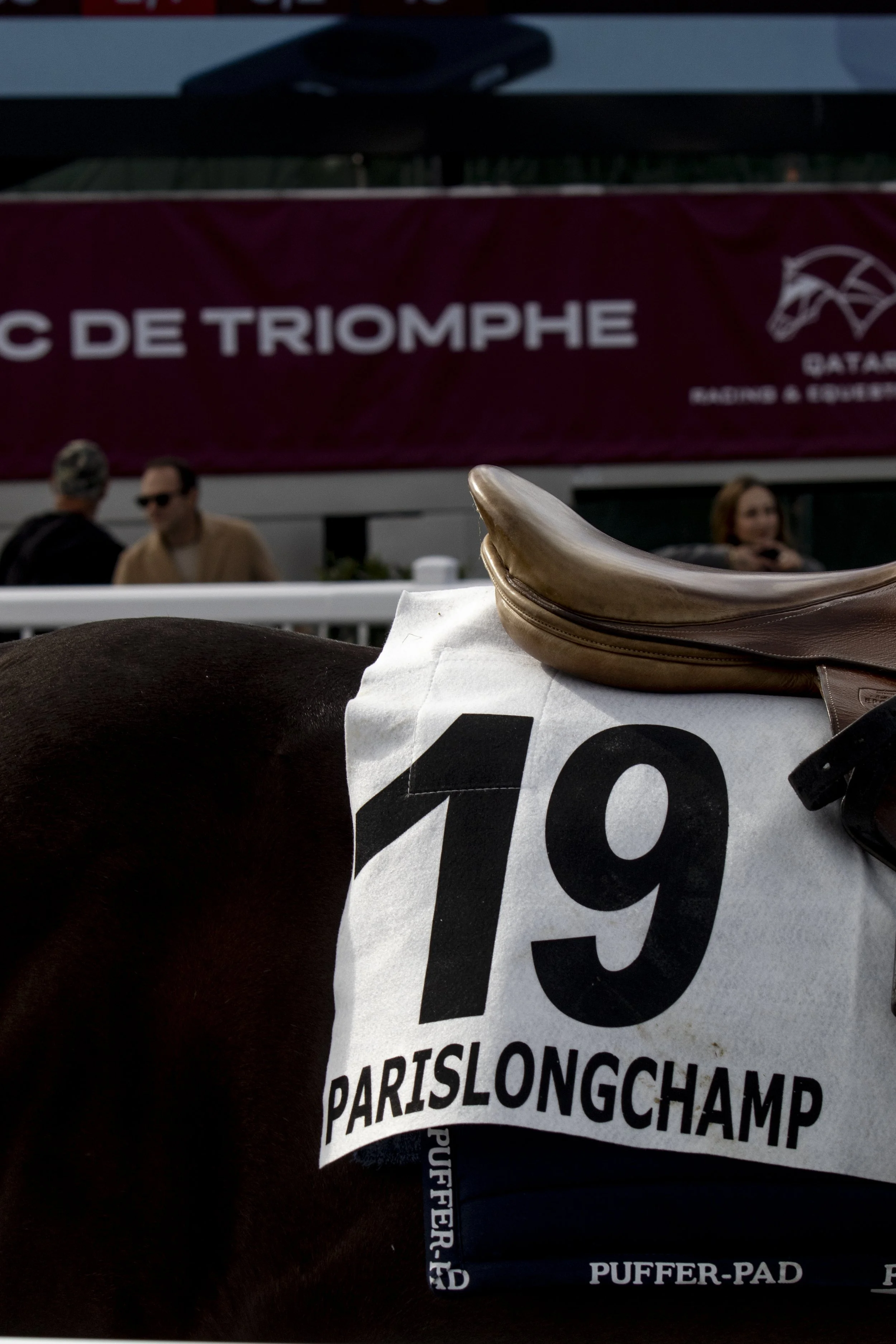 A racing event with horses and jockeys, with an advertisement banner in the background that reads 'C DE TRIOMPHE' and contains a logo for Qatar Racing & Equestrian Club.