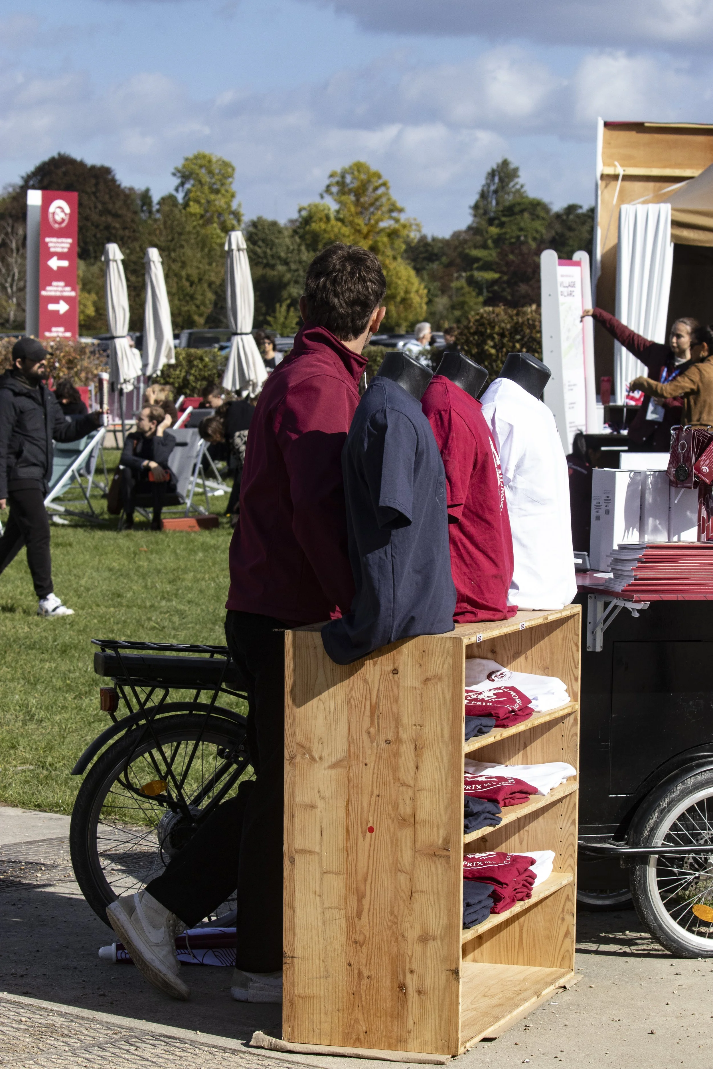 A person selling t-shirts from a wooden cart on a sunny day at an outdoor event, with people sitting and standing around, and trees in the background.