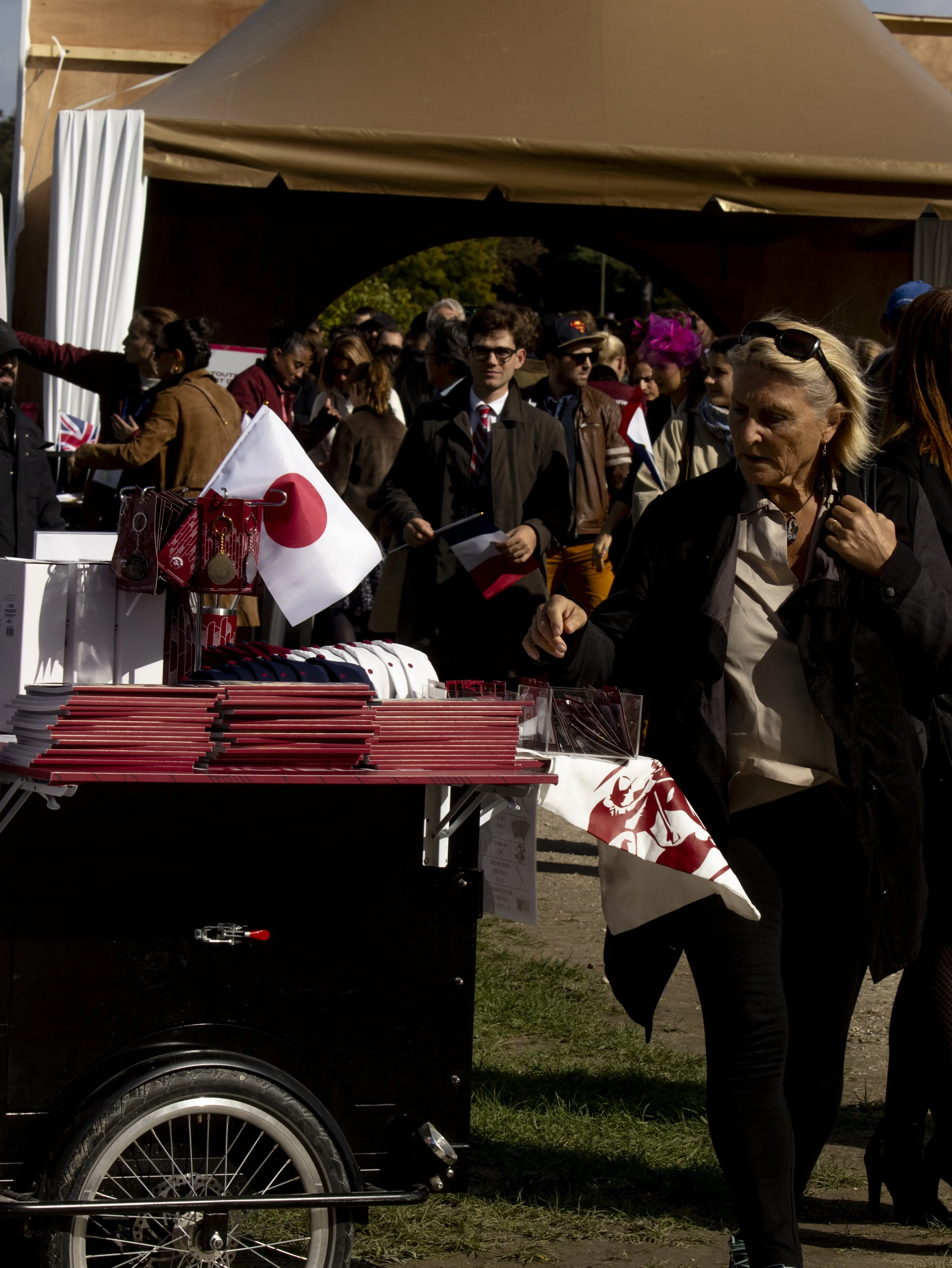 Crowd of people gathered under a large tent at an outdoor event, with a table in the foreground displaying various items, including a Japanese flag and red merchandise.