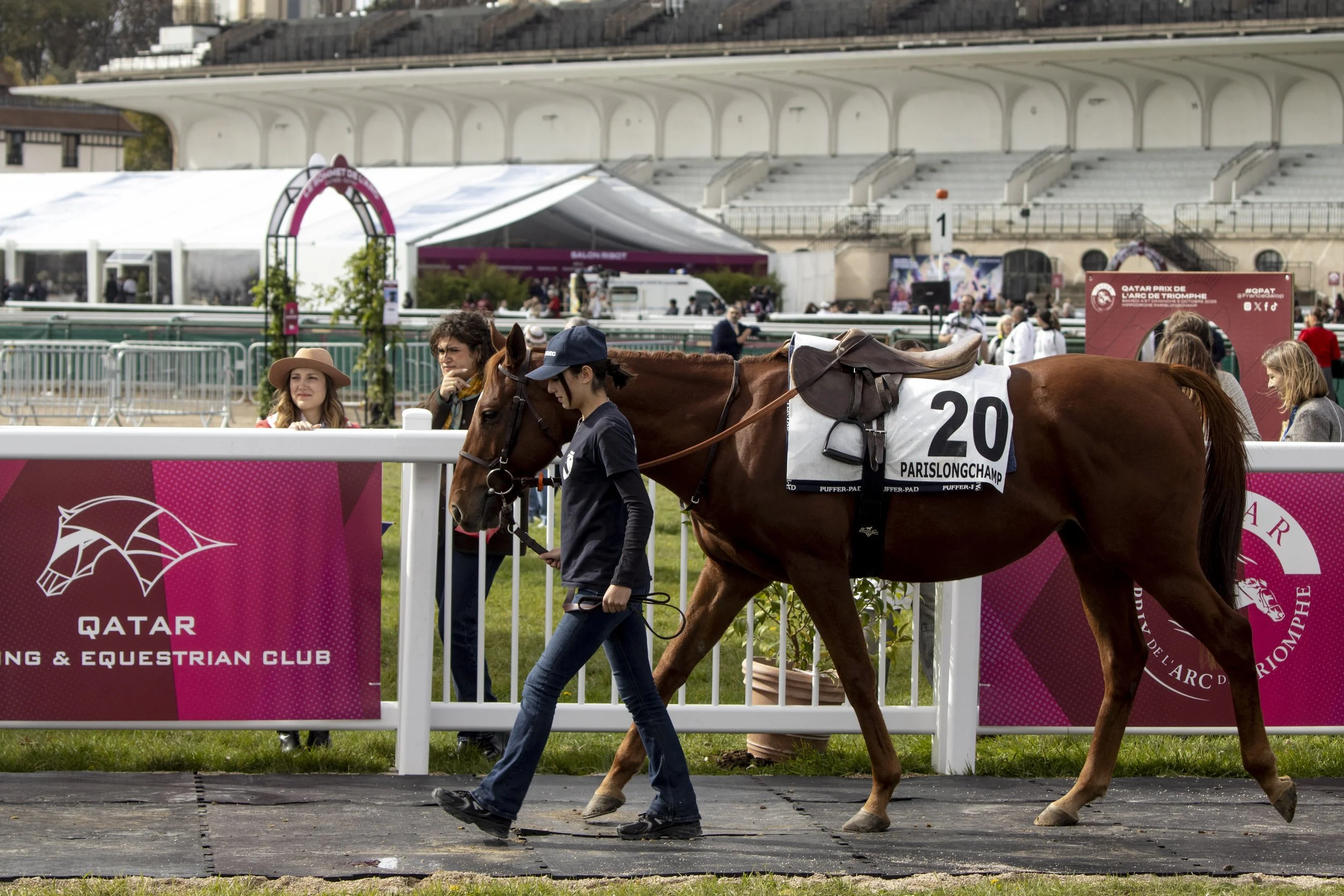A rider on a chestnut horse wearing a saddle, walking alongside a woman in a navy shirt and baseball cap holding the horse's lead at a horse racing event at the Qatar Prix de l'Arc de Triomphe. The background shows a grandstand and an event tent, wit