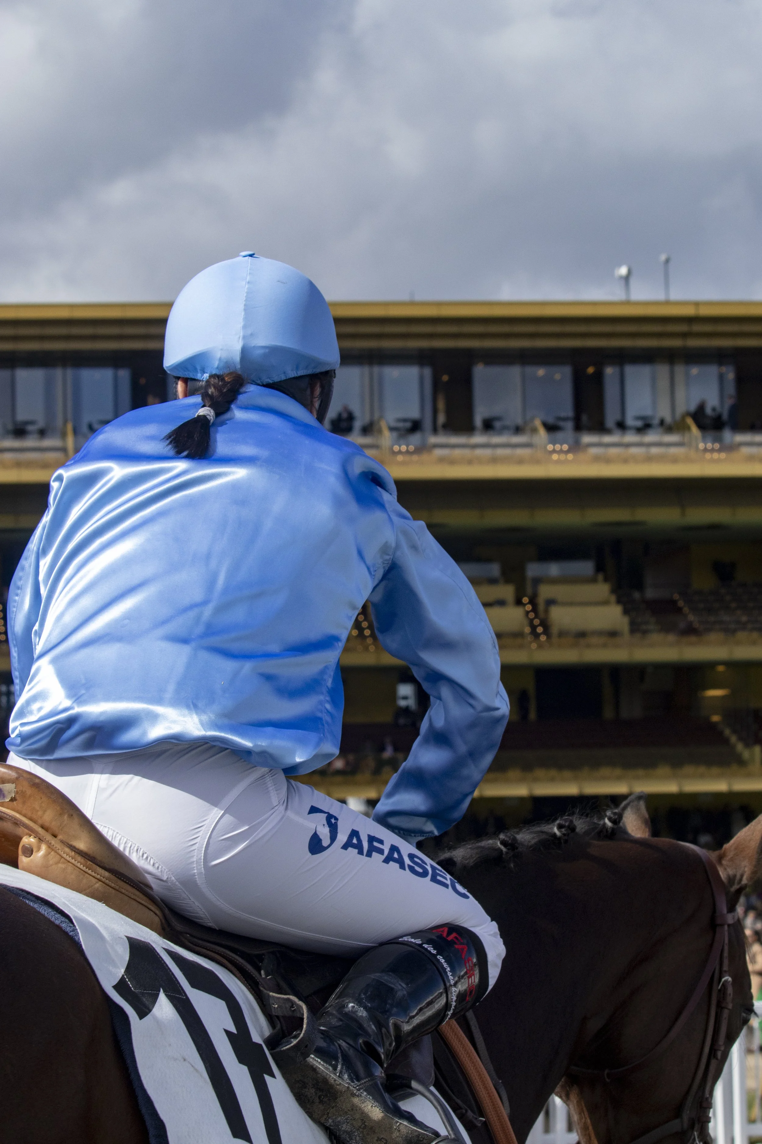 A jockey dressed in a light blue jacket and helmet, riding a dark brown horse with a white saddle pad marked 'AFAS'. They are at a racecourse, with possible spectators or facilities in the background under a cloudy sky.