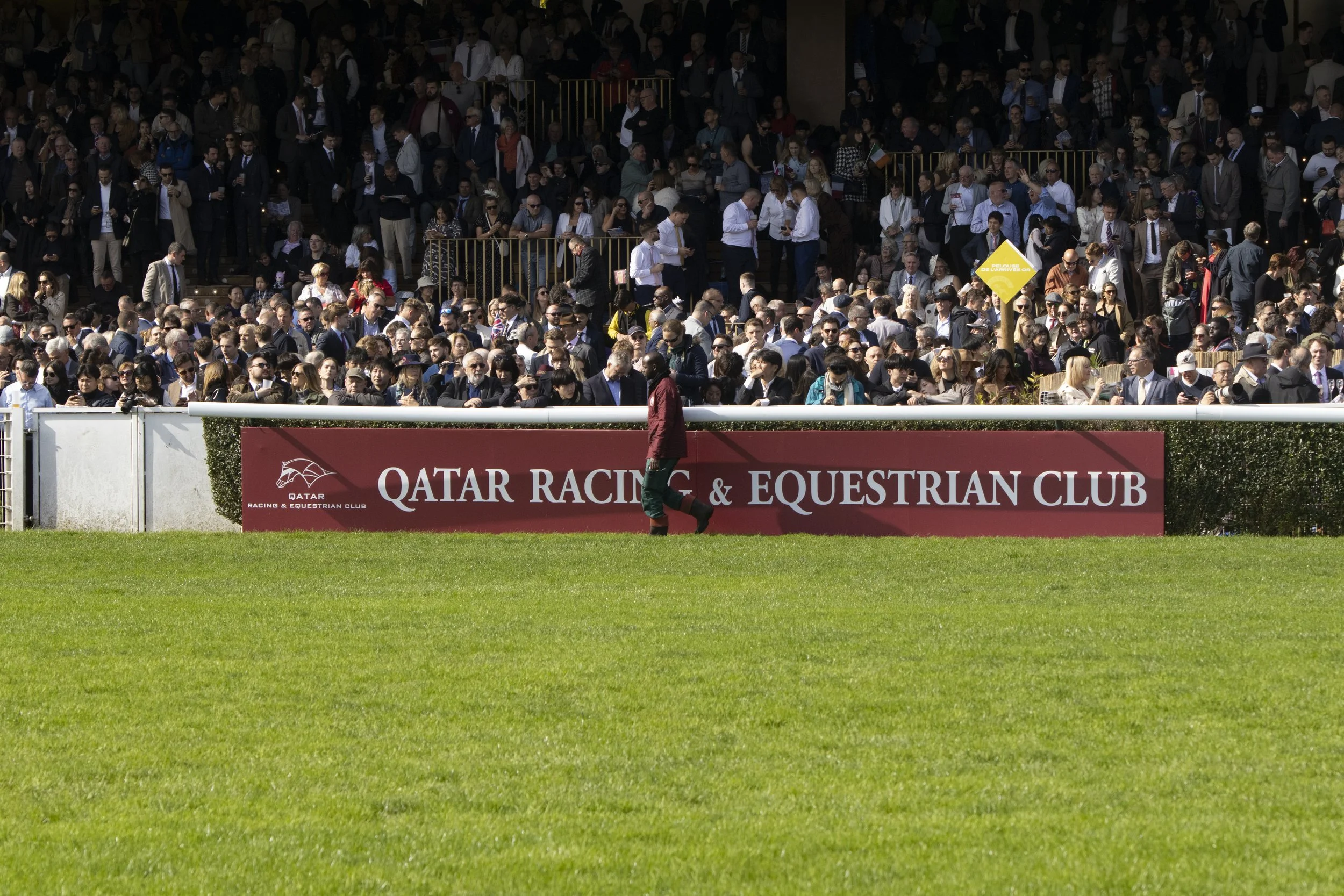 Crowd of people gathered at the Qatar Racing & Equestrian Club, some standing and some sitting, with a person walking in the foreground, and a sign indicating the club's name on a red barrier at the bottom of the image.