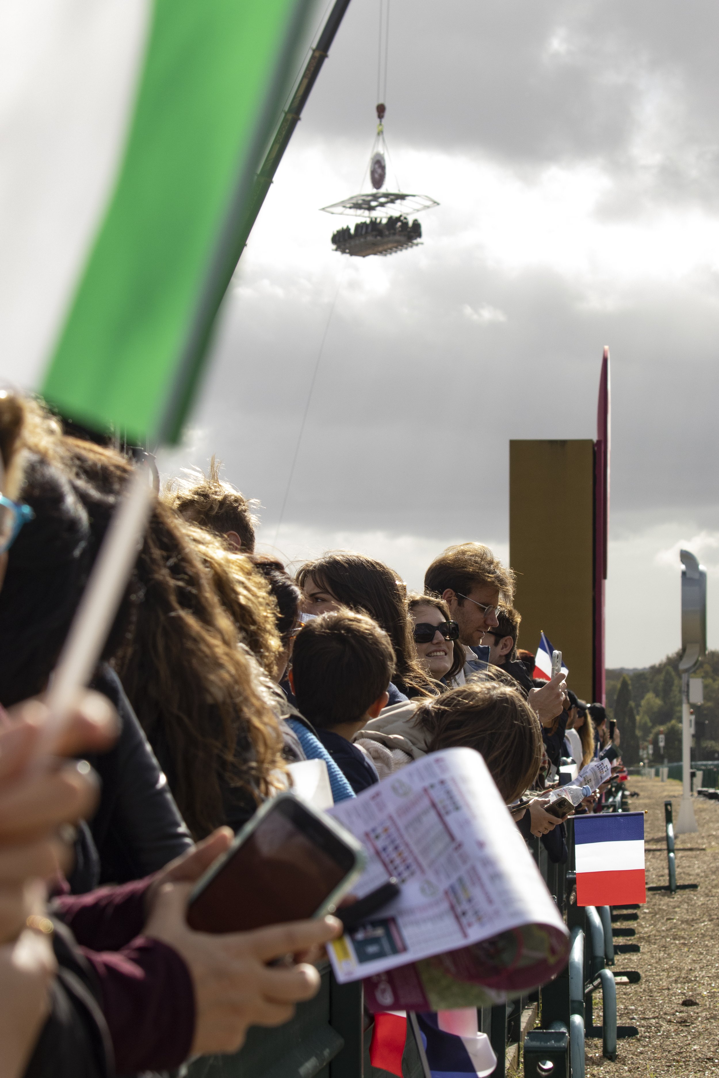Crowd of people standing behind a barrier, some holding flags and pamphlets, with overcast sky and clouds in the background, at an outdoor event.