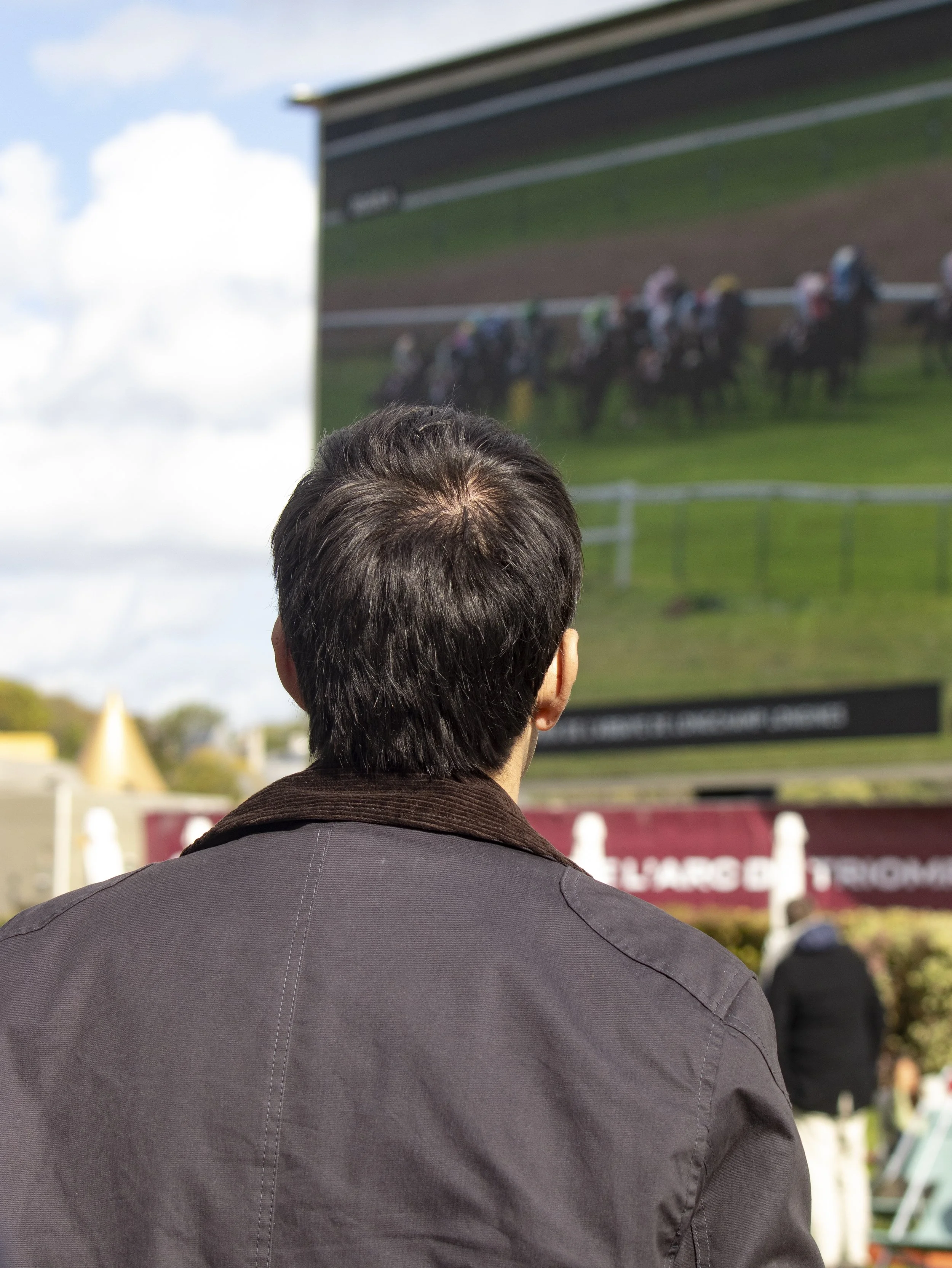 A person with dark hair wearing a gray jacket watching an outdoor horse race on a large screen.