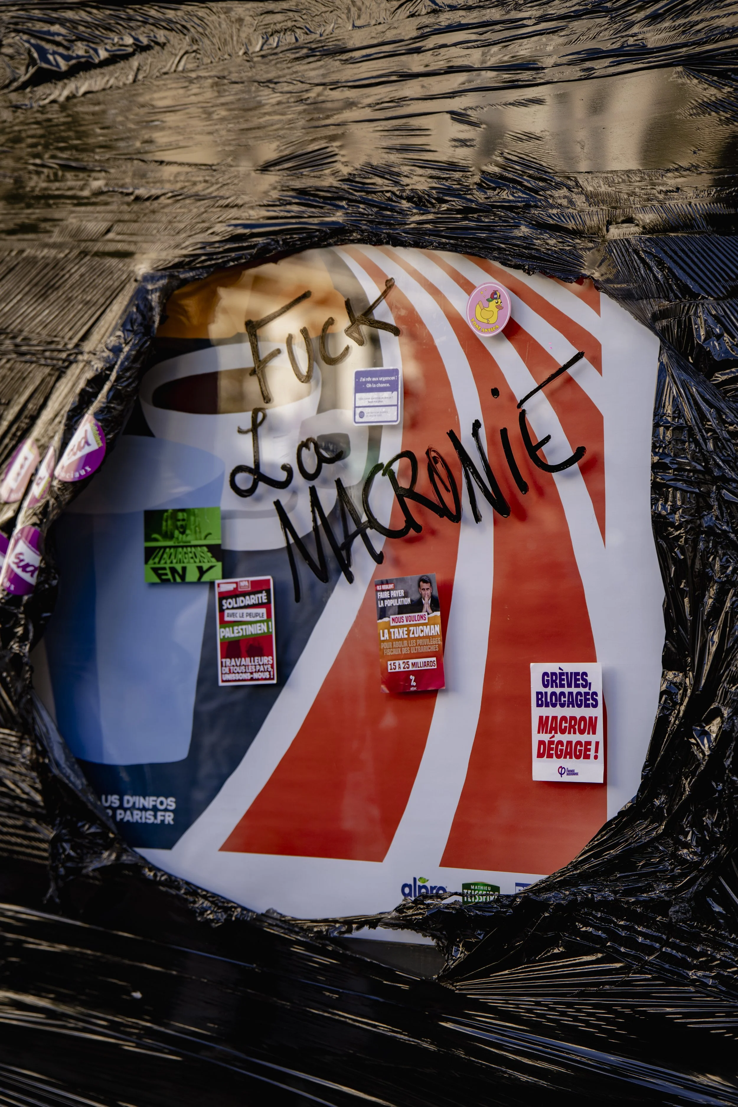 A poster with handwritten words 'FUCK LA MACRON' over a cup of coffee and various stickers, partially covered with black plastic wrap.