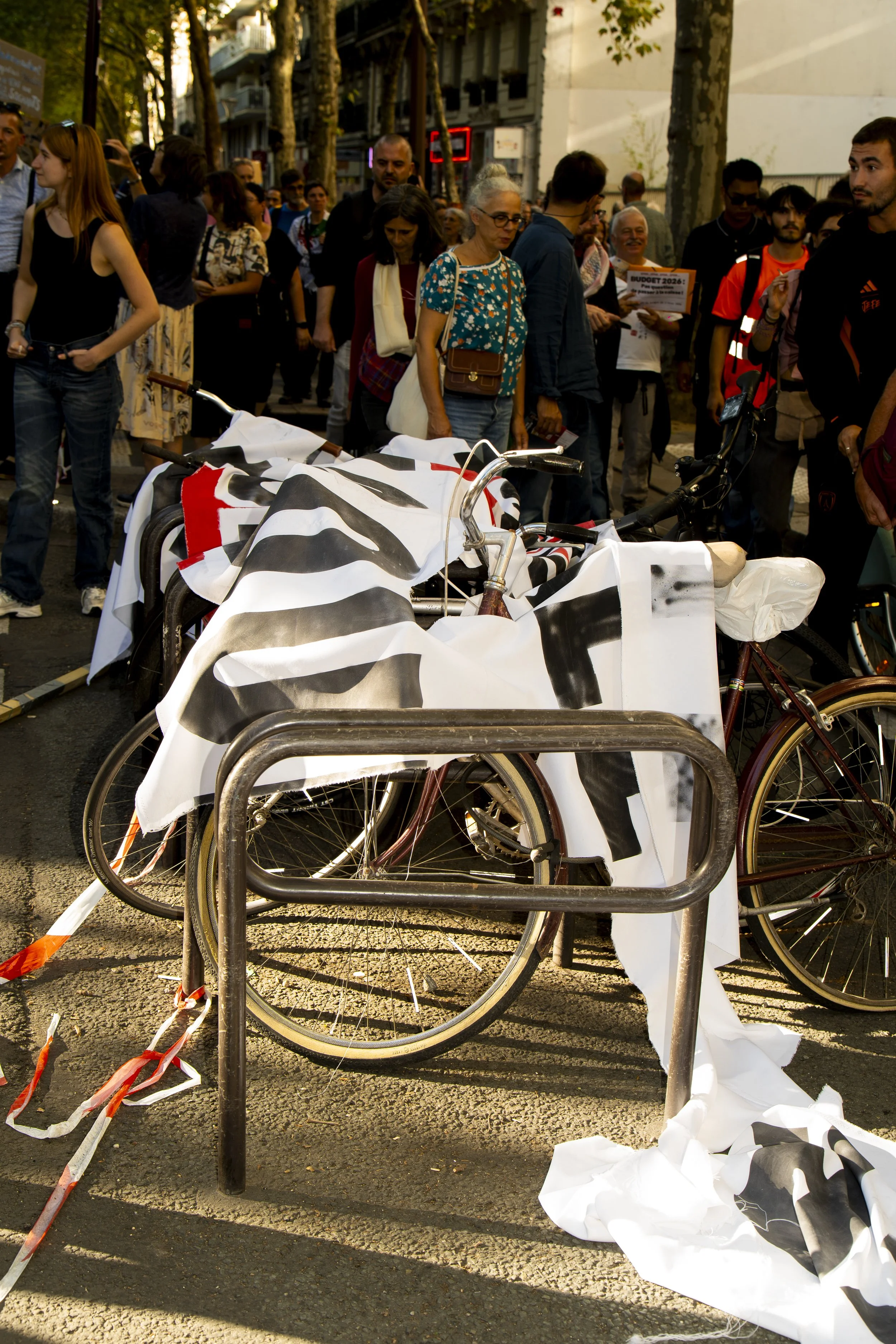 Several bicycles are locked to a metal bike rack, with some having black and white flags draped over them. In the background, a crowd of people is walking along a tree-lined street in an urban area.