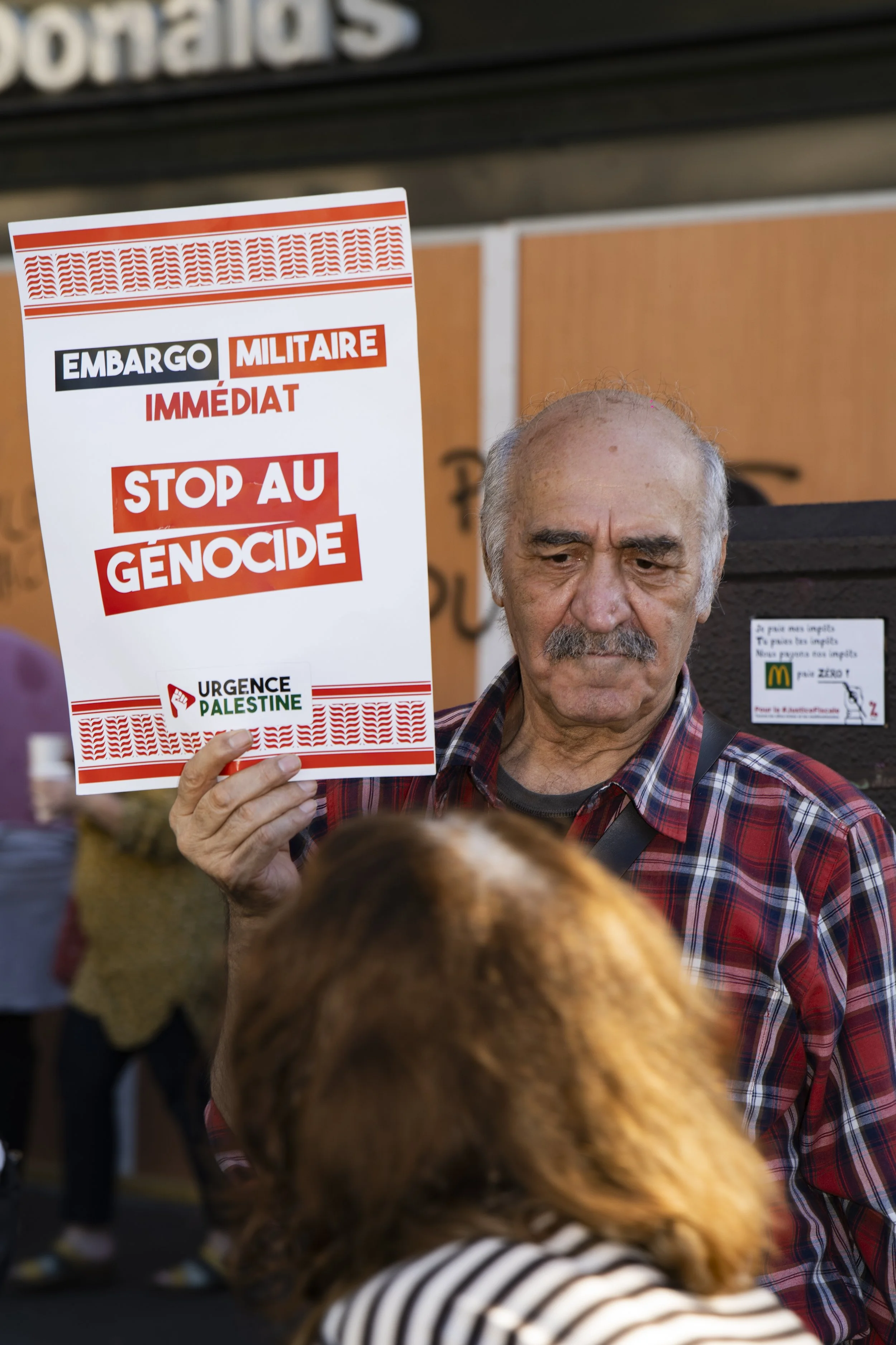 Man holding a protest sign that says 'Stop Genocide,' with various banners and people in the background at a demonstration.