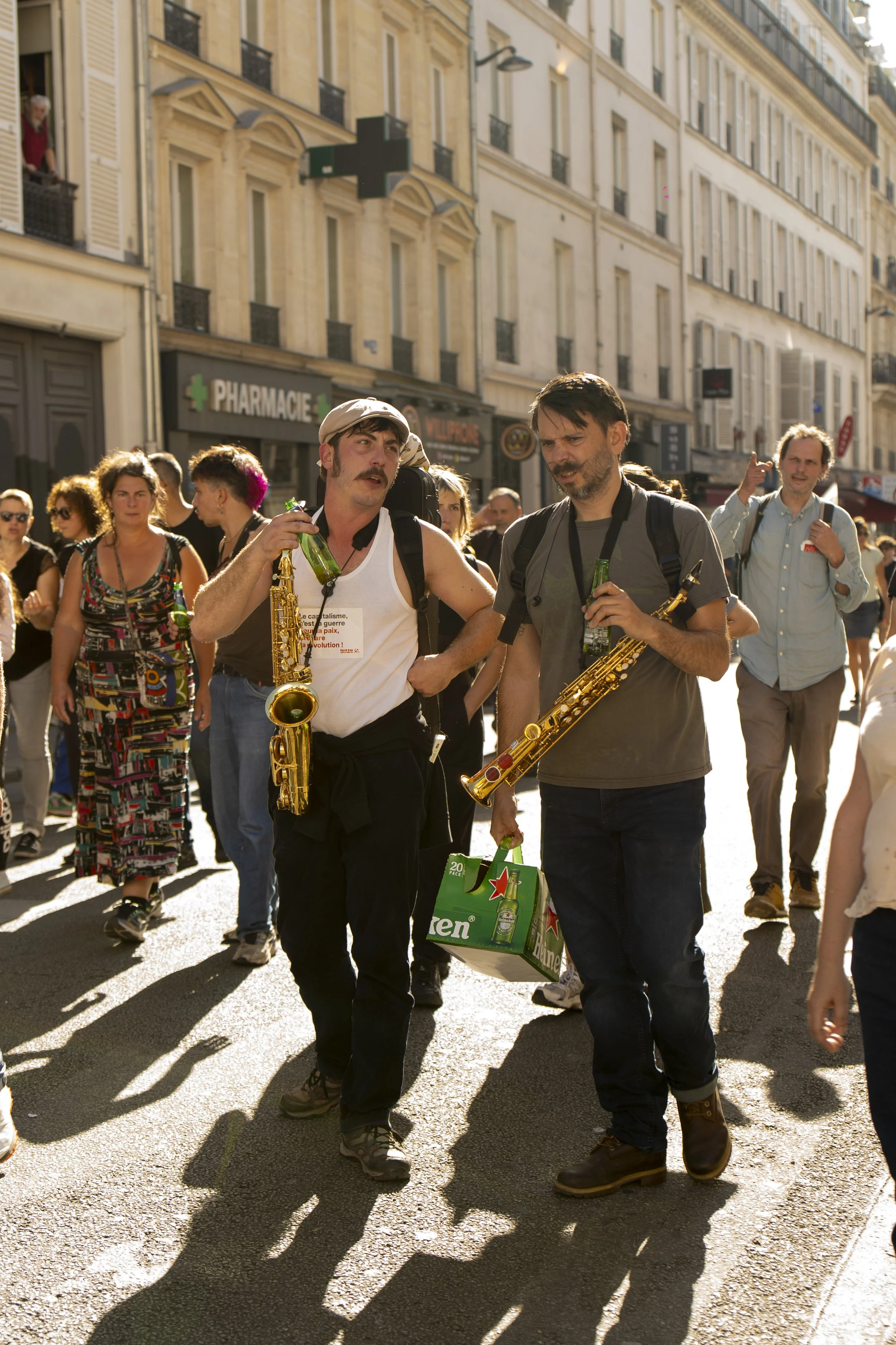 People walking on a sunny city street with buildings, including a pharmacy, in the background. Two men in the foreground are holding bottles and a pack of Heineken beer.