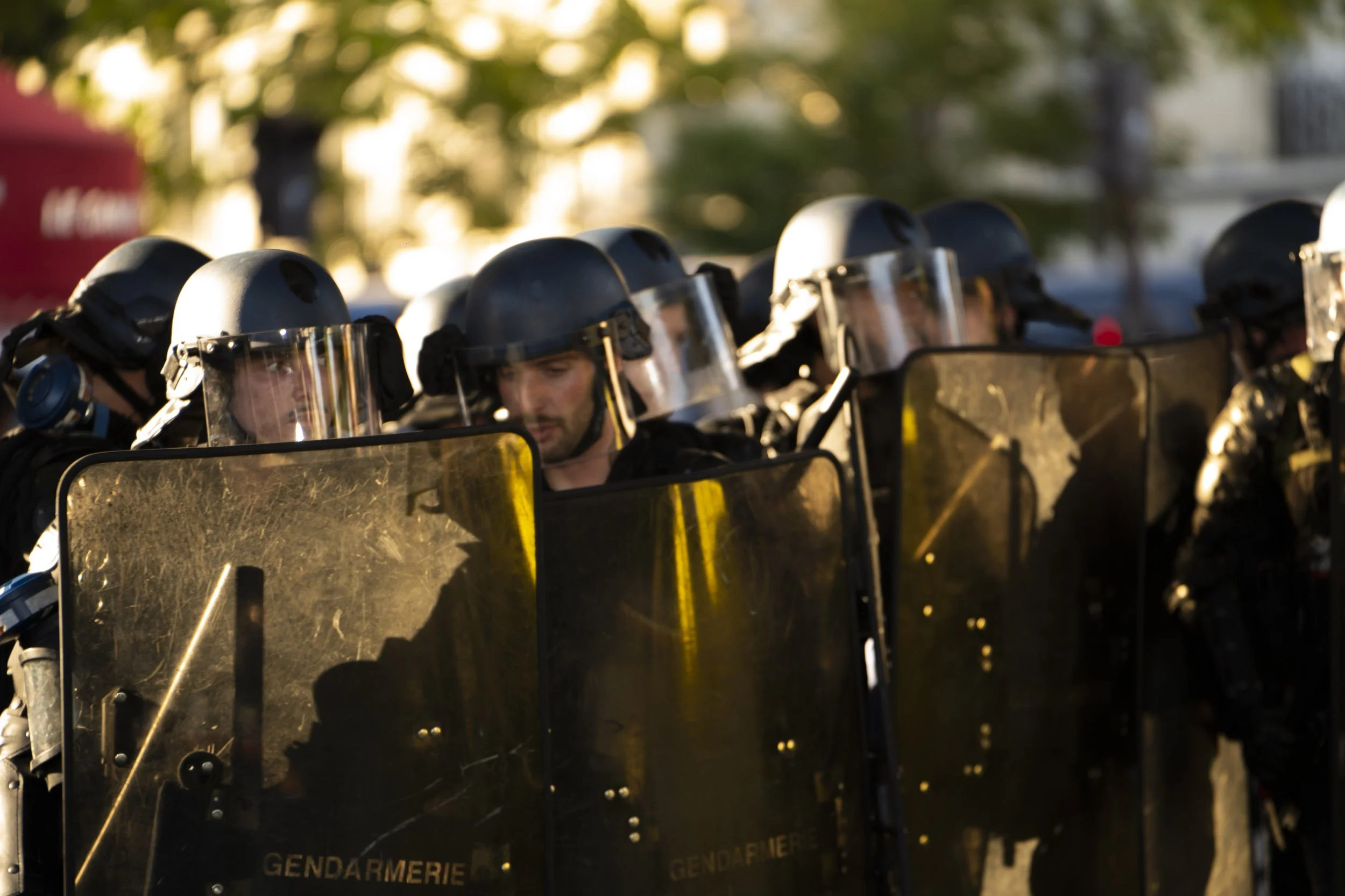 Line of riot police officers in helmets and shields standing outdoors, with trees and buildings in the background.
