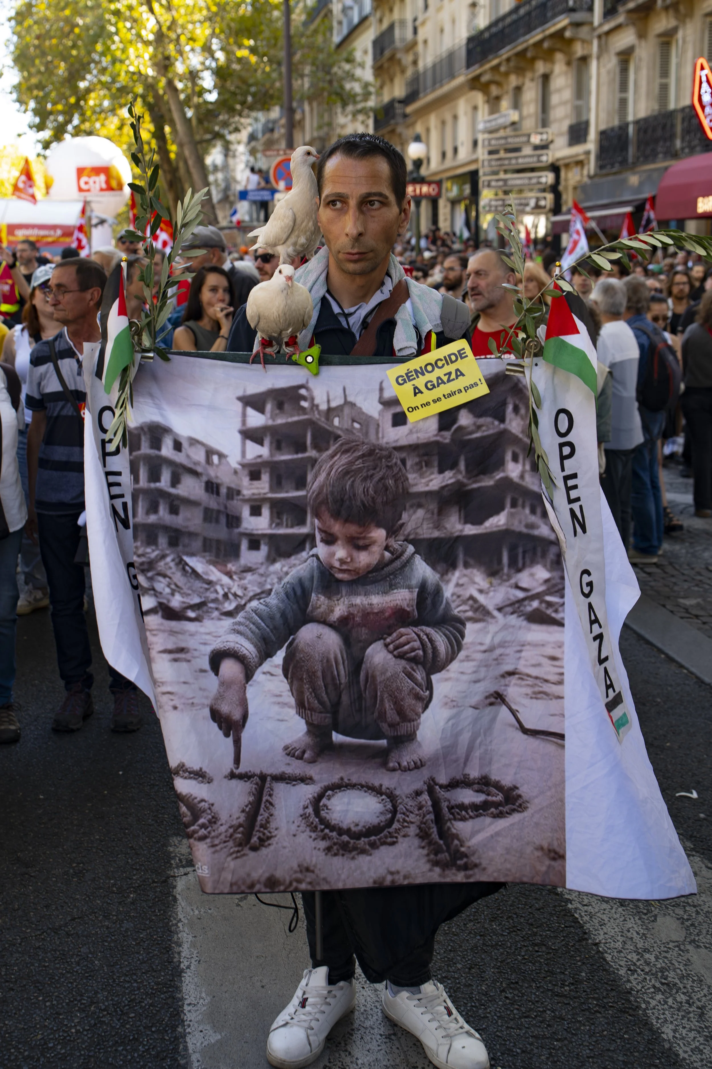 A man participating in a protest march holding a banner with an image of a child playing in the sand, with a destroyed building in the background. The man has a dove perched on his shoulder. There are other protesters behind him, and the scene is set