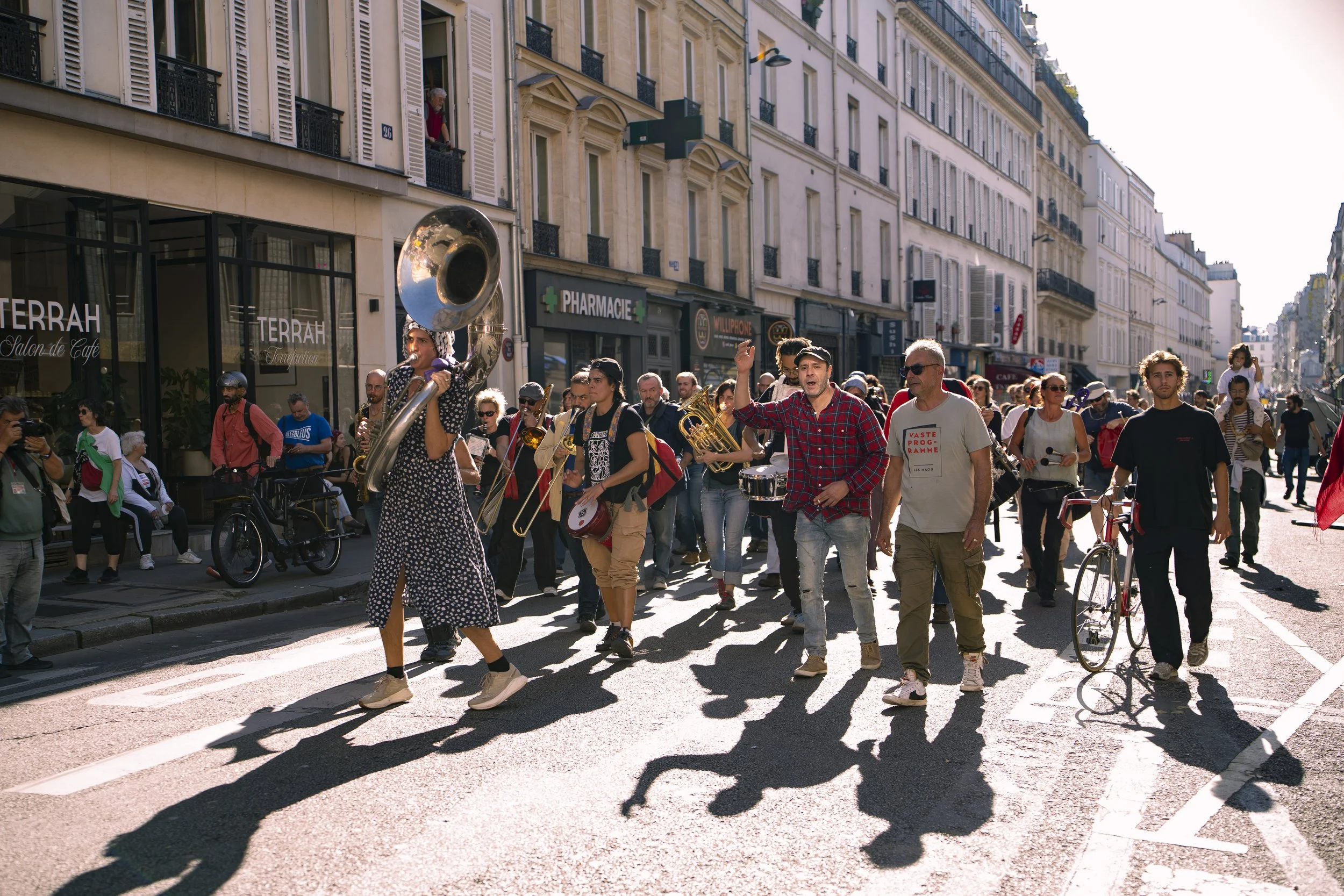 Musicians marching on a city street, playing instruments including a sousaphone and trumpets, with a crowd of pedestrians watching, in front of cafes and pharmacies, in bright daylight.