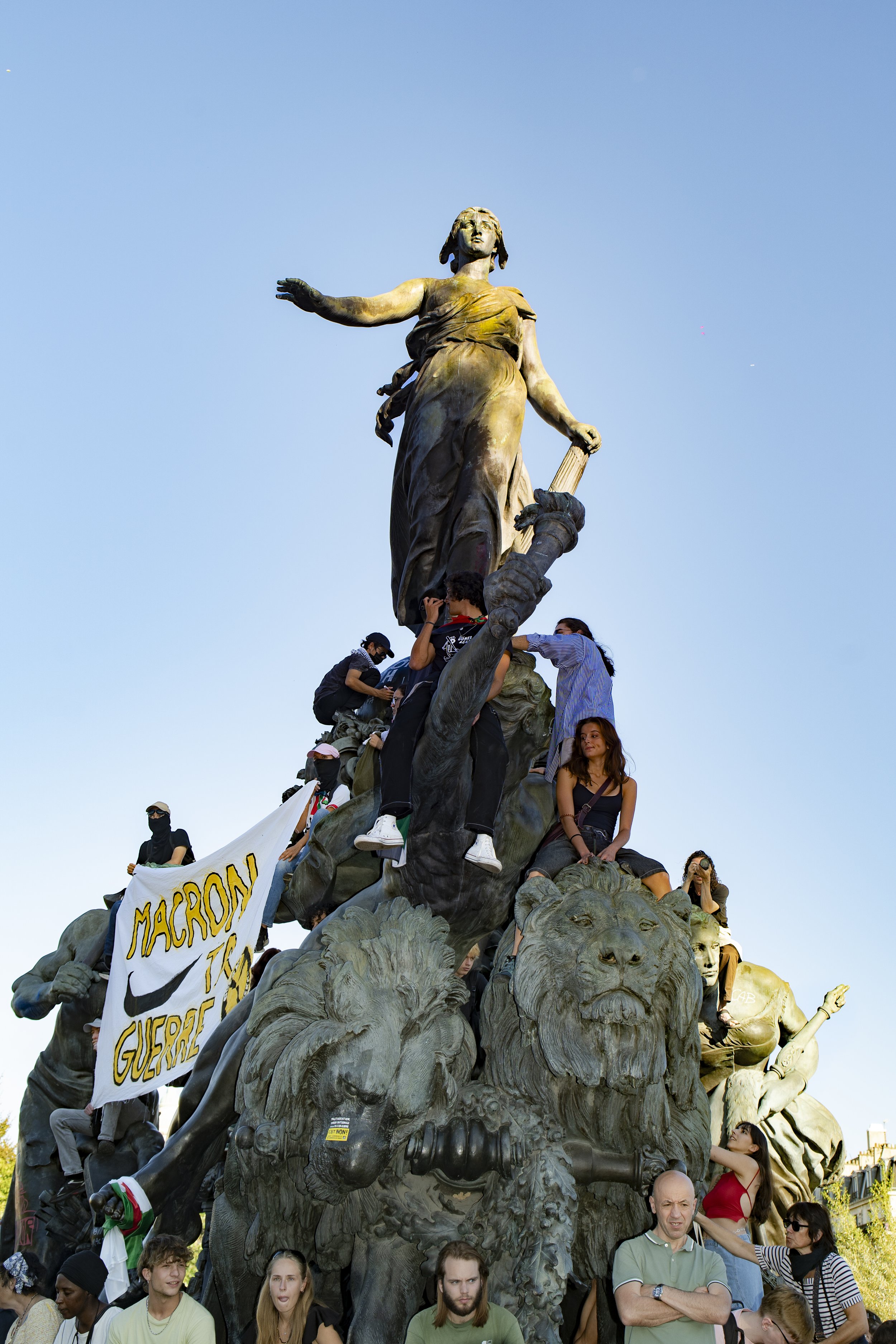 People protesting on a large statue with a flag and banners, some public figures are visible.
