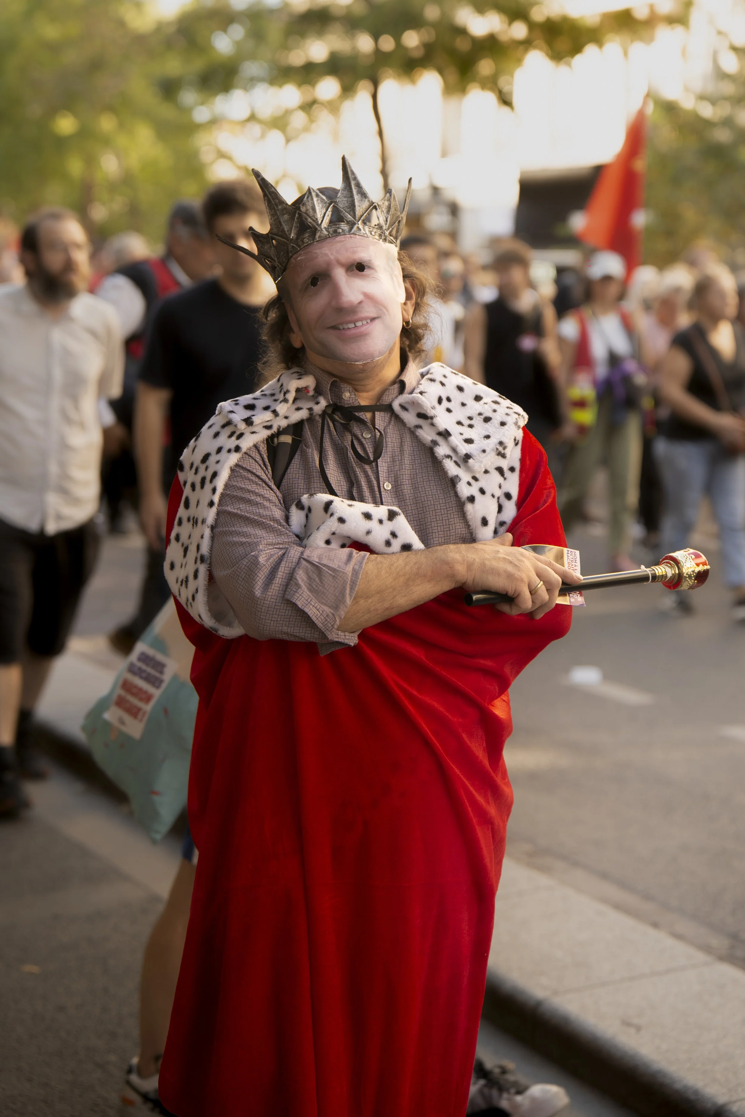 Man dressed as a king in a red robe and crown, standing with arms crossed, at a gathering or event outdoors, with a group of people in the background.