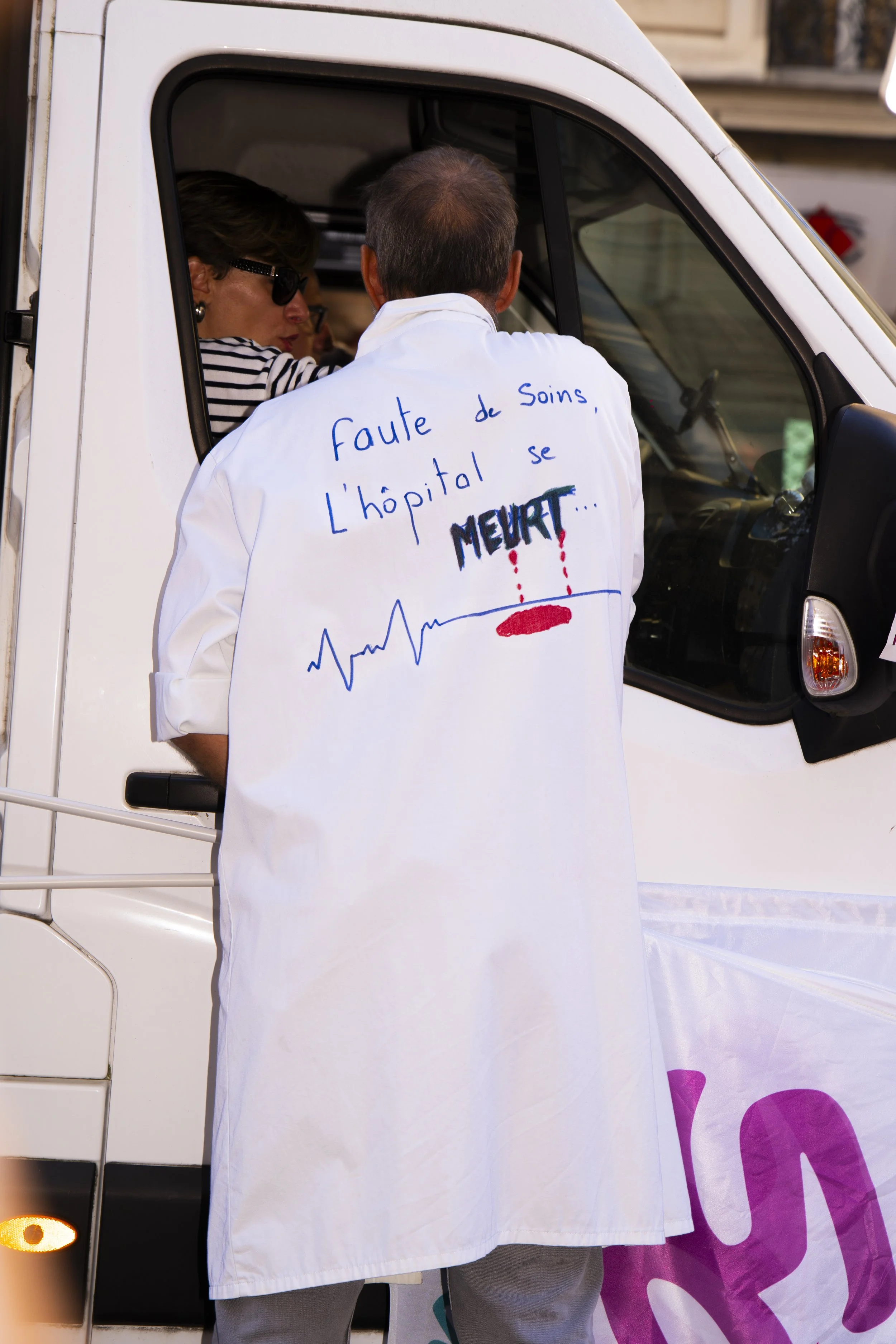 A man wearing a white coat with French writing and bloodstains on the back, talking to a woman inside a small white vehicle, possibly at an event or demonstration.