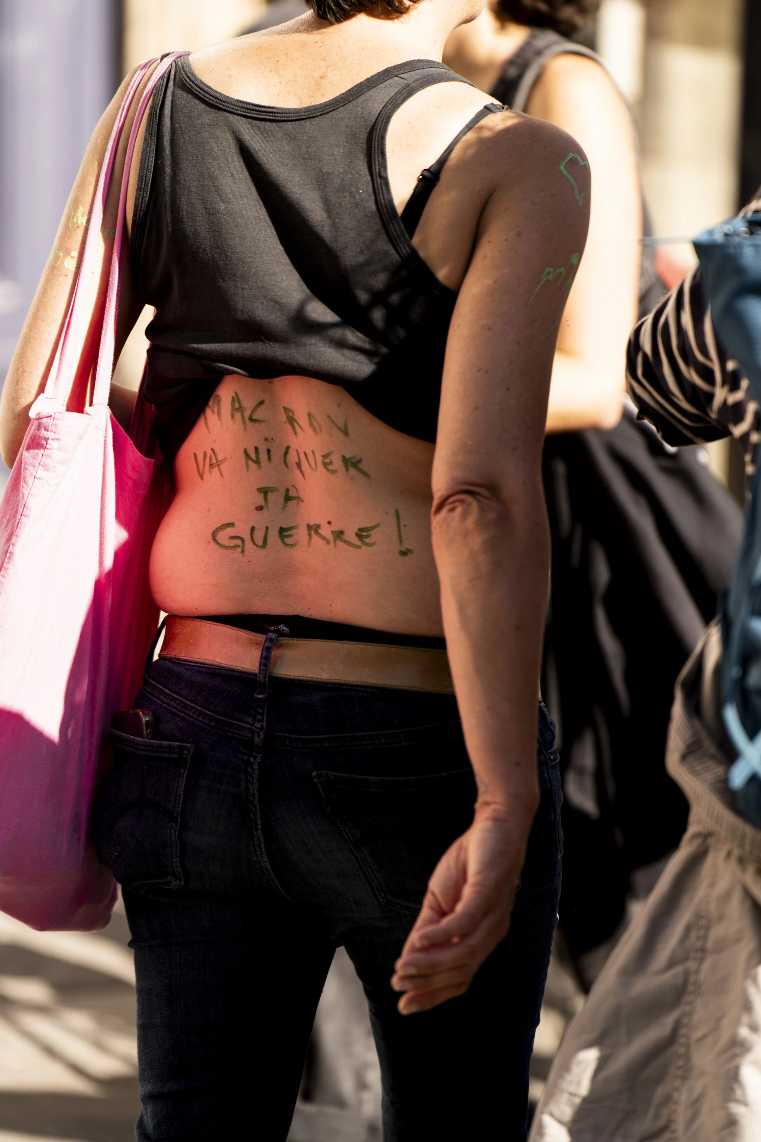 Close-up of a woman with a temporary tattoo on her shoulder and writing on her side, wearing a black tank top and jeans, standing among other people.