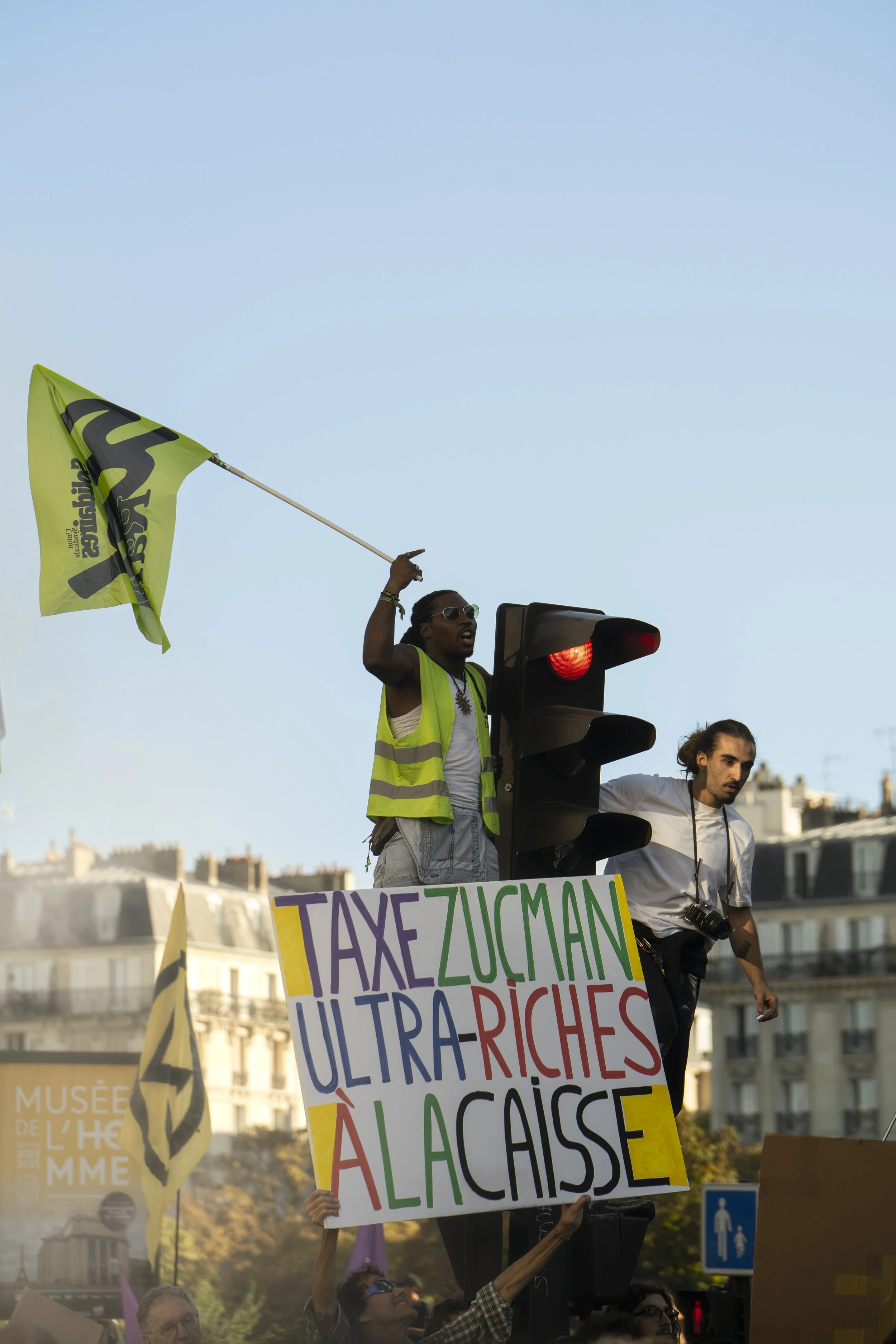 A protest scene in an urban area with people holding colorful signs and banners, and one person waving a yellow flag with black text. The background shows buildings and trees, and the protest appears to focus on social or political issues.