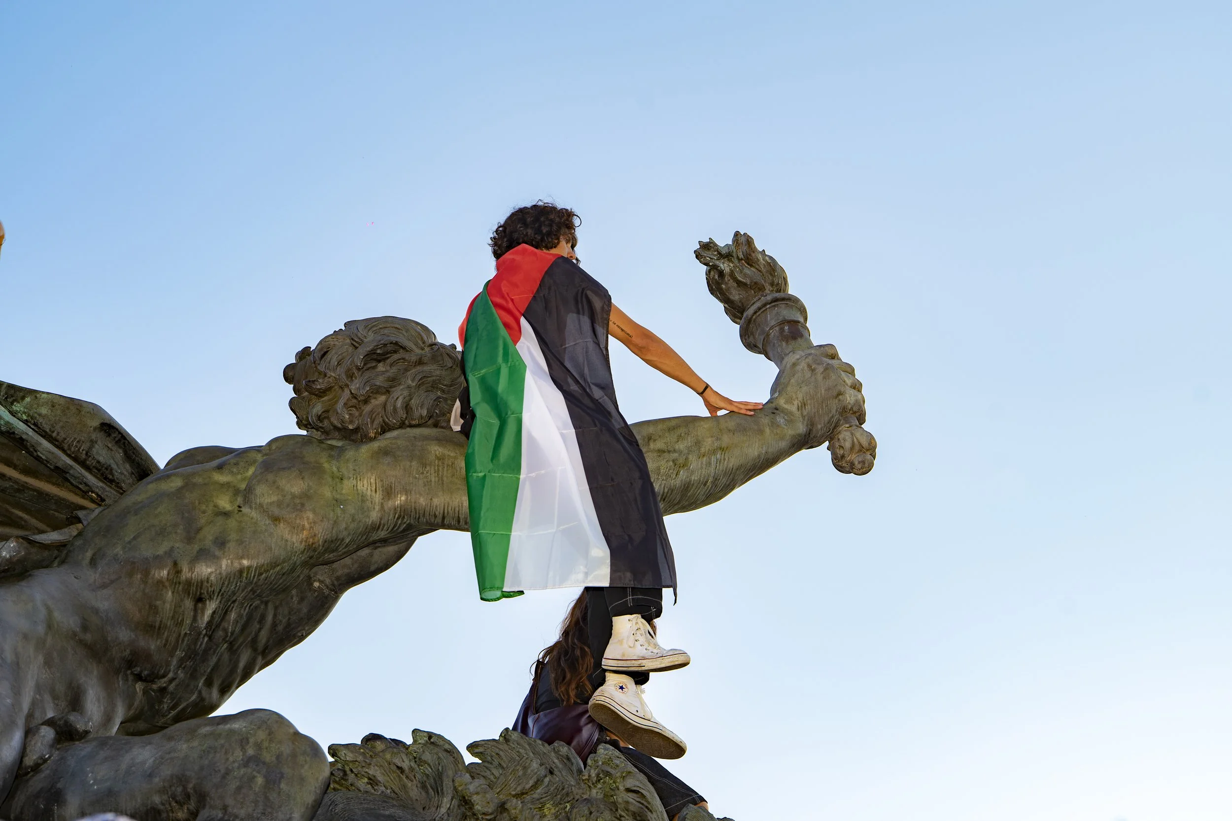 People climbing on a large bronze statue with a blue sky in the background. One person is sitting on the statue's arm, draped in a Palestinian flag.