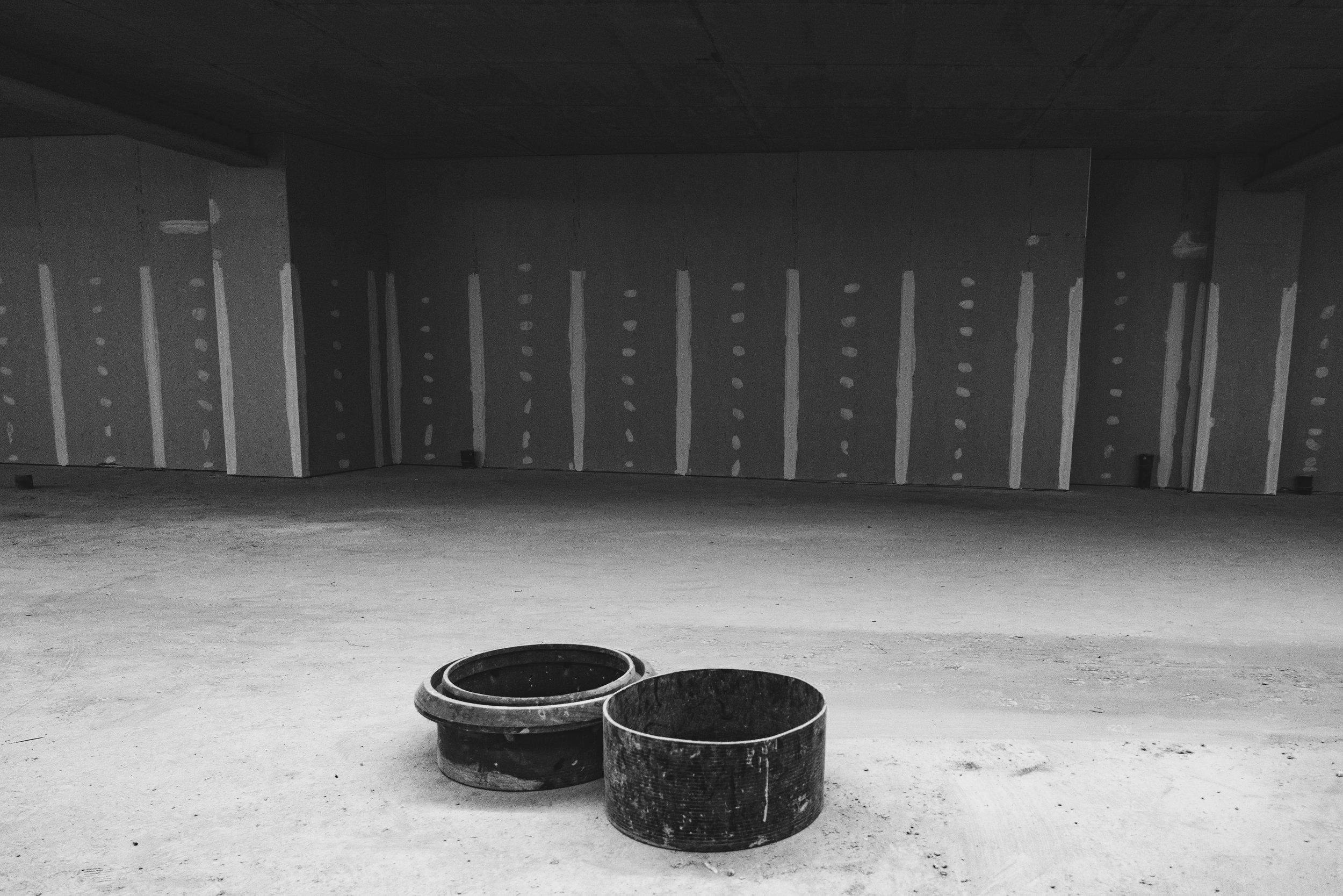 Underground parking garage with unfinished concrete walls and floor, large circular openings in the wall, and two black spray-painted metal buckets on the floor.