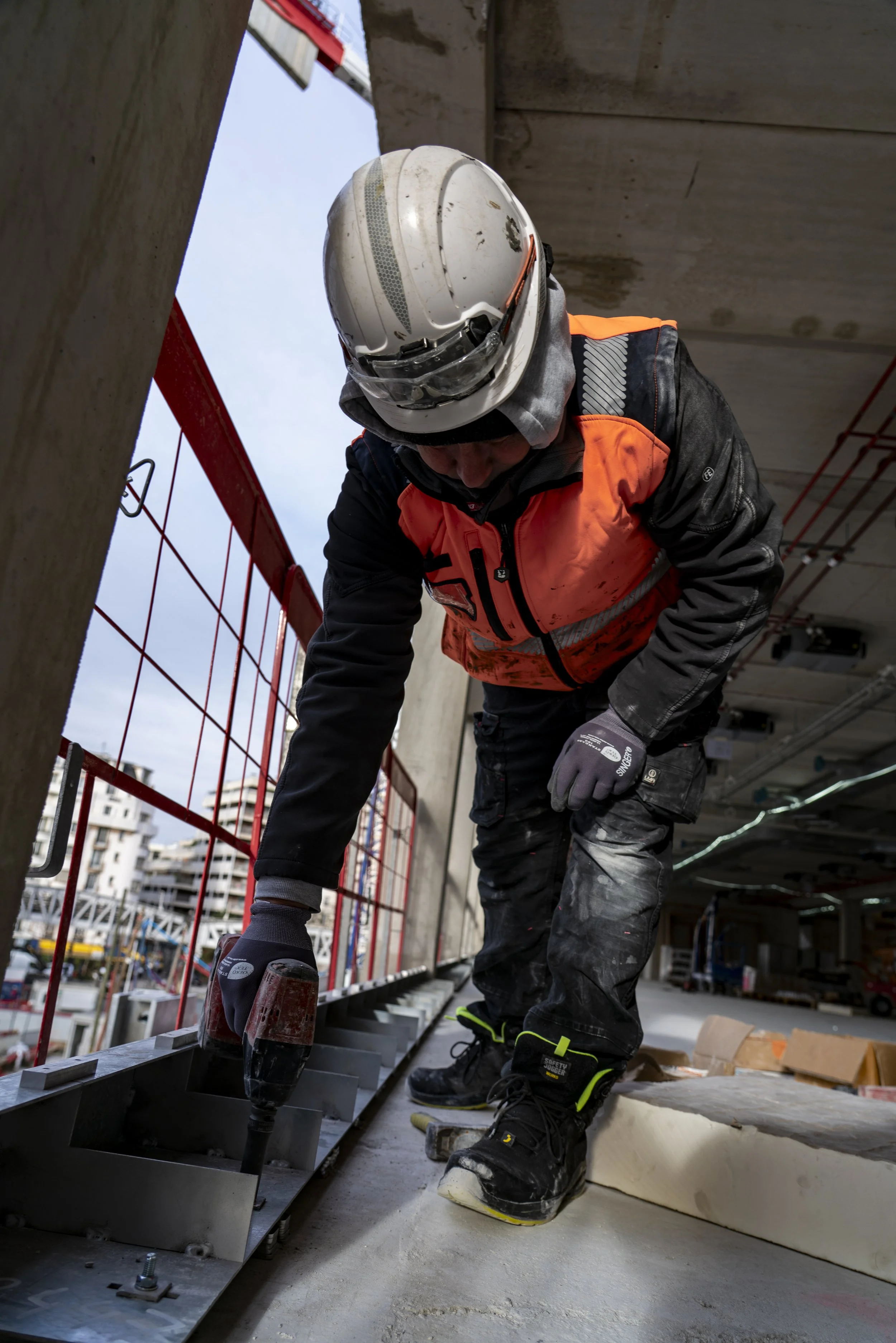 Construction worker wearing a white safety helmet and orange safety vest working on steel framework on a construction site. He is also wearing gloves and sturdy boots, and the background shows city buildings and an overcast sky.