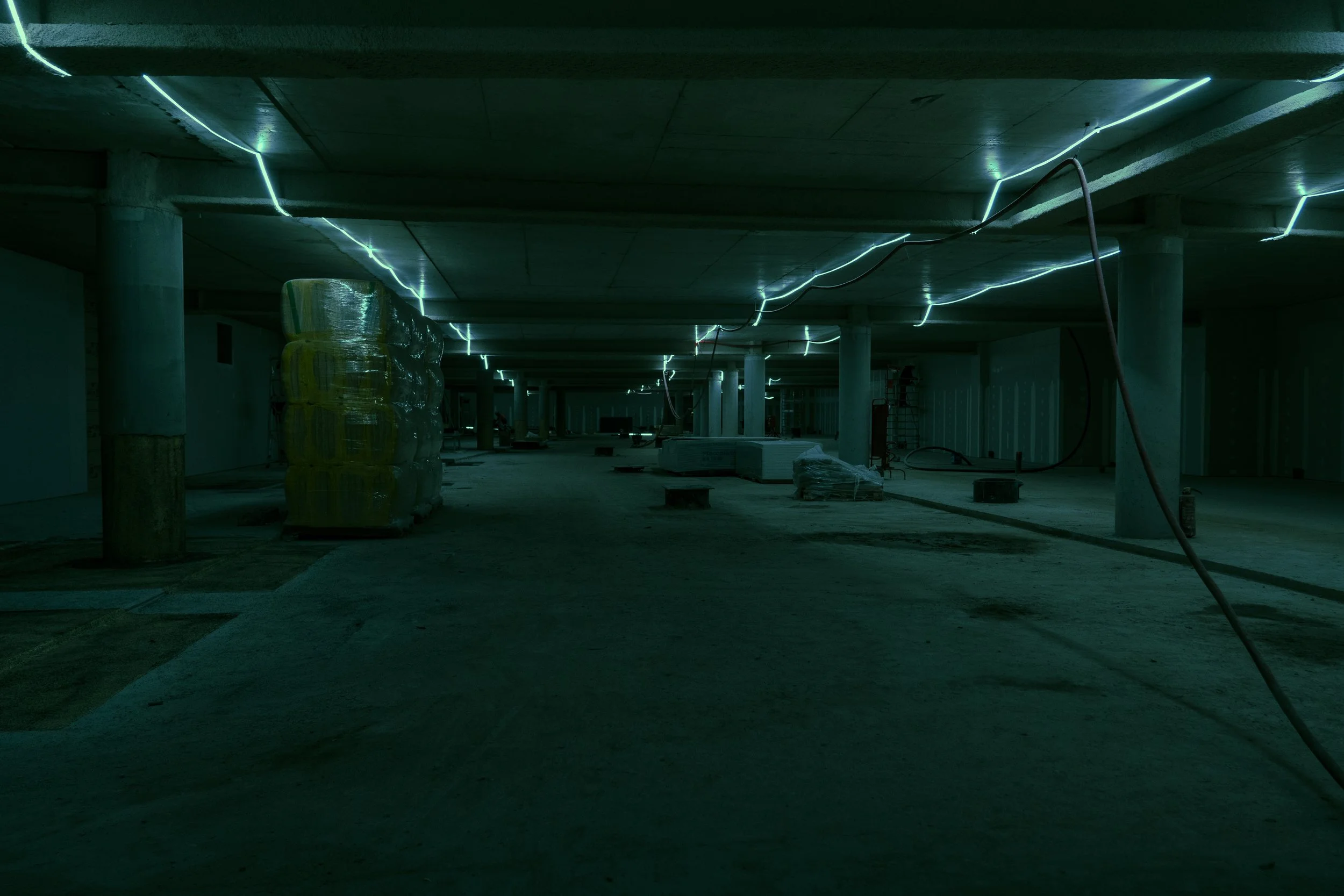 Underground parking garage or construction site with concrete pillars, low ceiling, and blue-green LED strip lighting on ceiling and support beams.