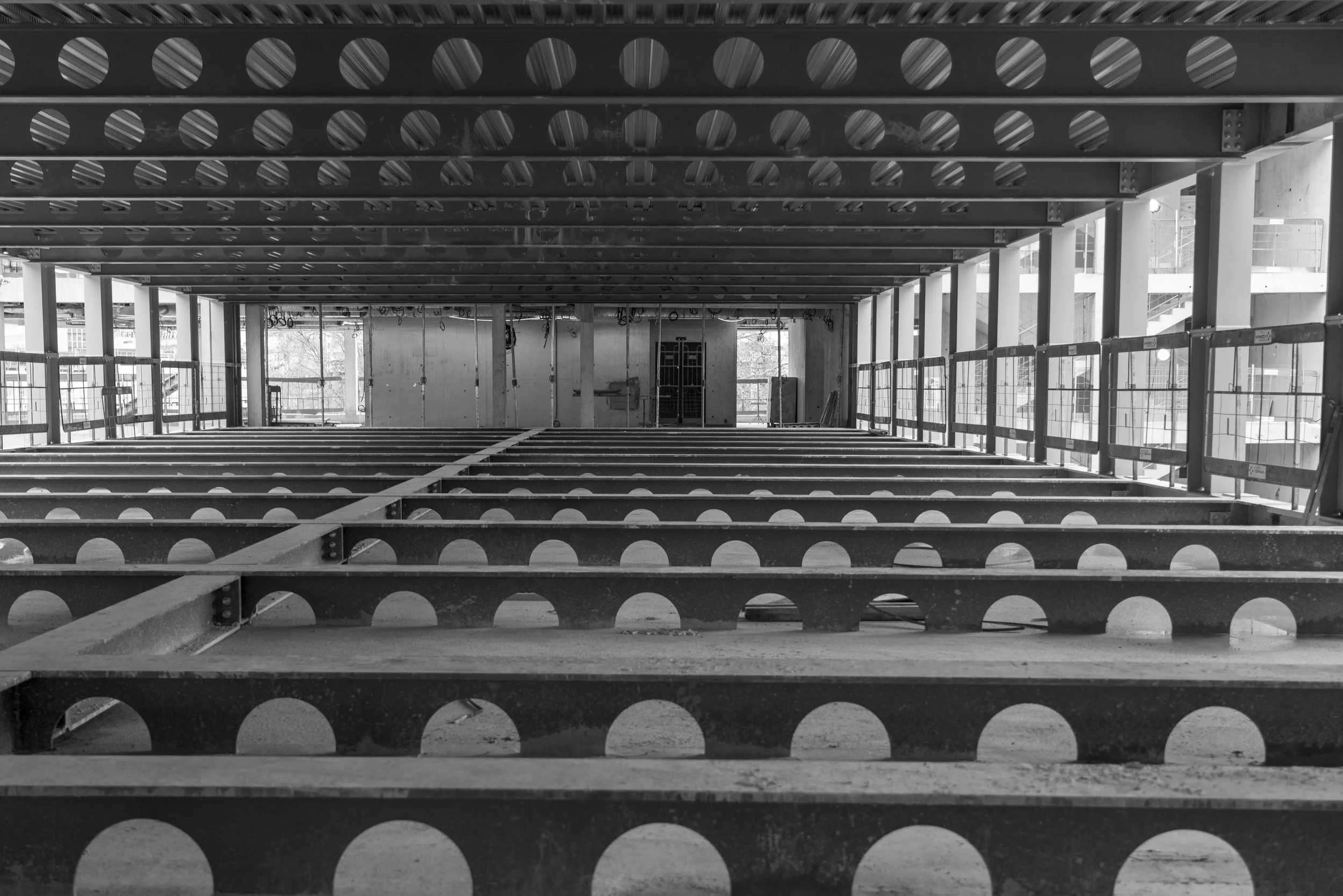 Black and white photo of a construction site with metal beams and circular cutouts, with scaffolding and an unfinished building structure in the background.