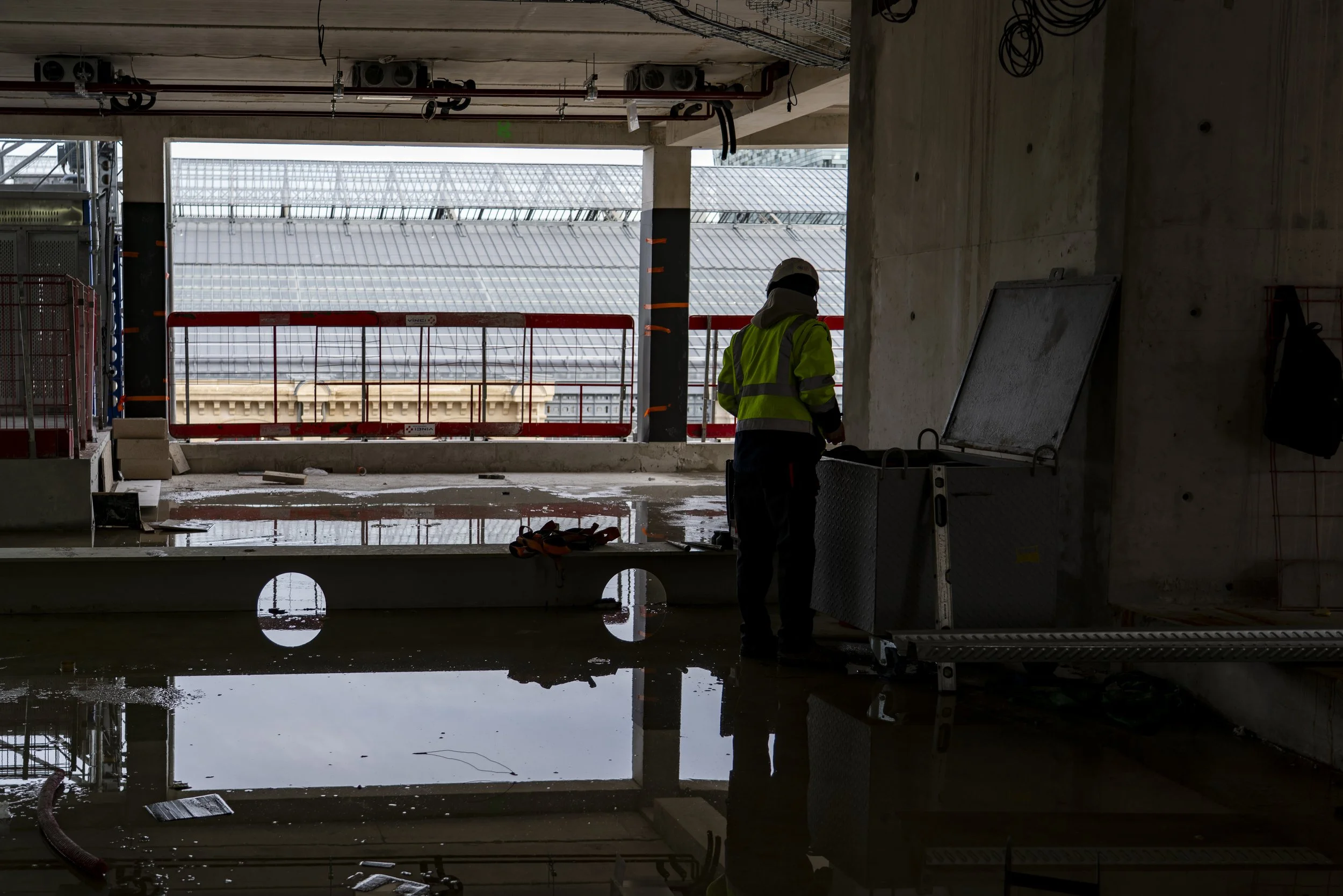 Construction worker wearing a safety helmet and reflective vest standing inside a building under construction, with a large window opening and scaffolding outside, reflected in a puddle of water on the floor.