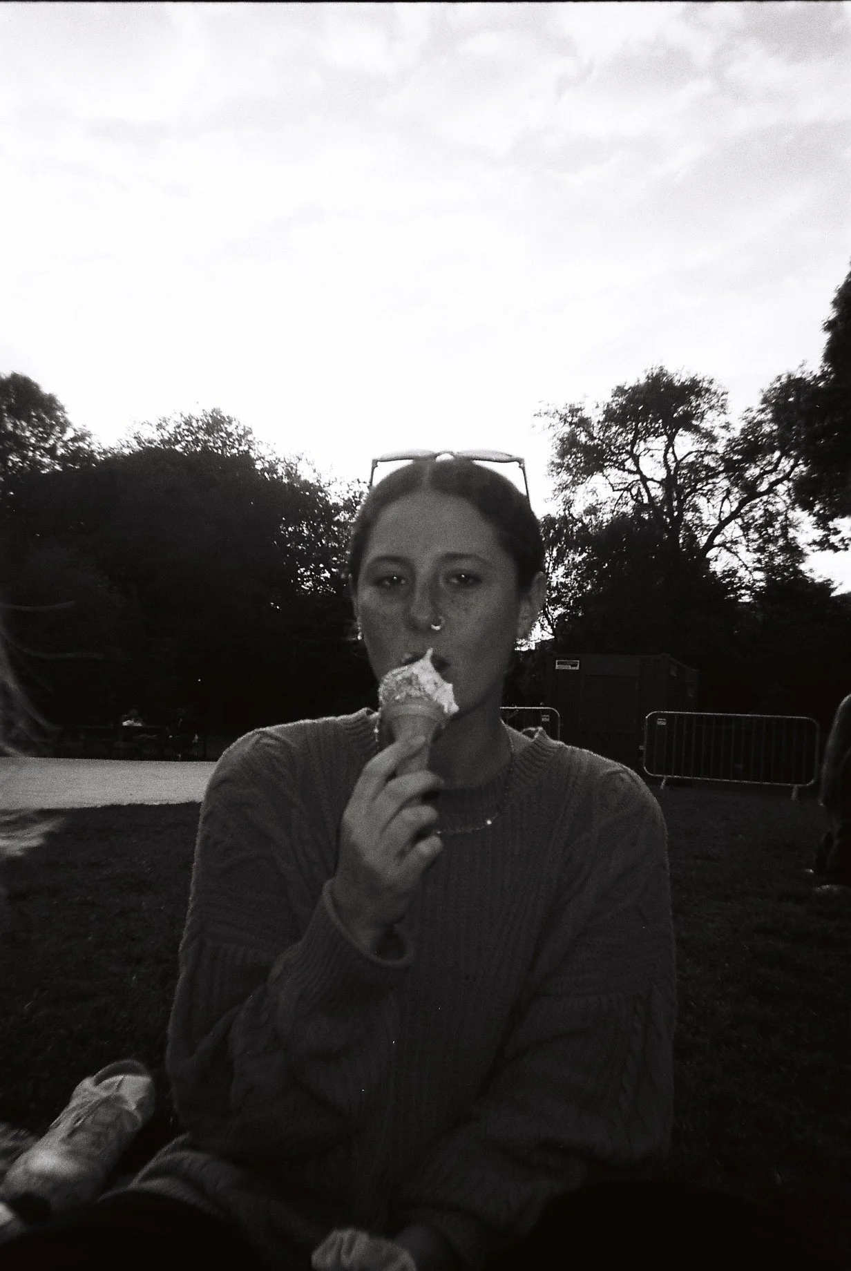 A woman with sunglasses on her head eating ice cream outdoors, with trees and a barrier in the background.