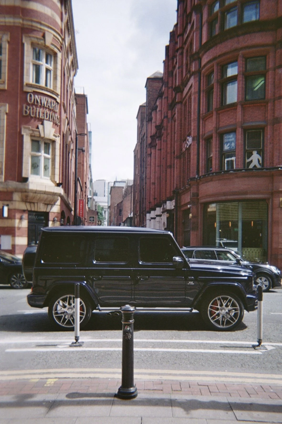 A city street scene with red brick buildings, parked cars, and a black luxury SUV in the foreground, with street posts and crosswalk lines visible.